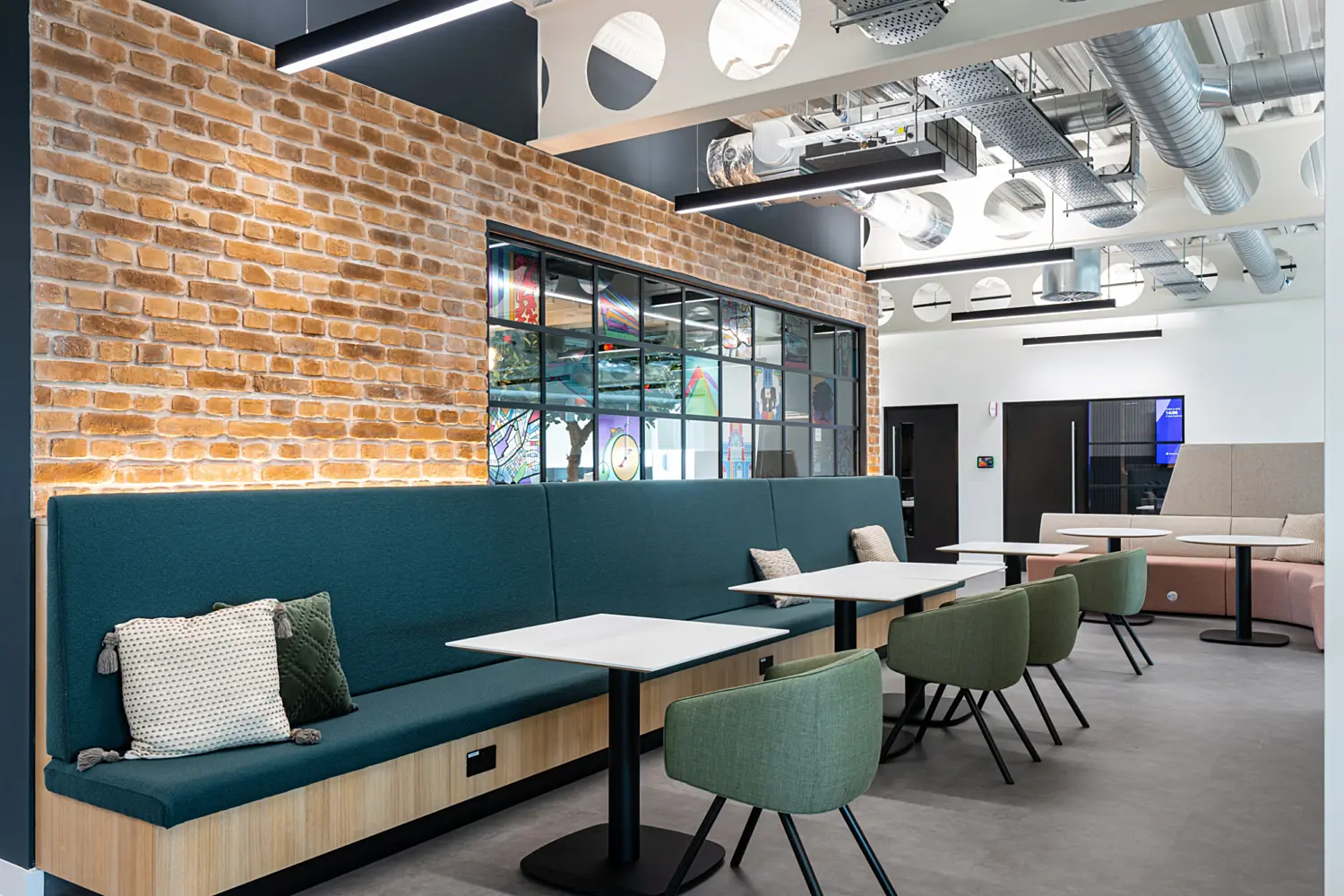 A modern office lounge with exposed brick and white walls, featuring a long teal upholstered bench paired with small white tables and green chairs. The ceiling includes exposed ductwork and circular cutouts, adding a contemporary touch. A secondary seating area with beige upholstery is visible in the background.