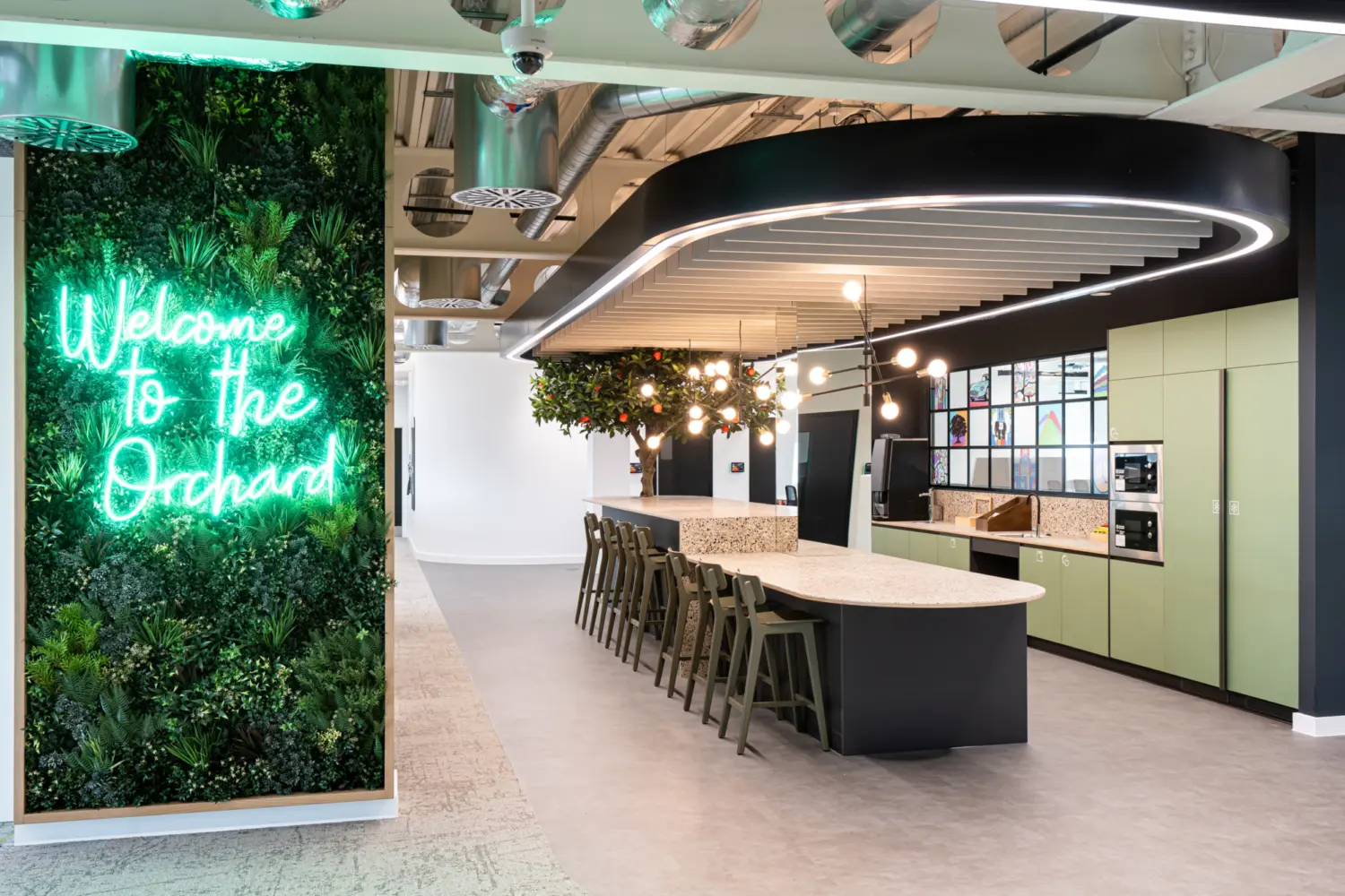 A stylish modern office kitchen with a long island counter, bar stools and overhead lighting. A vertical garden wall on the left displays neon text reading "Welcome to the Orchard." Wall mounted cabinets and clean finishes complete the inviting, contemporary workspace.