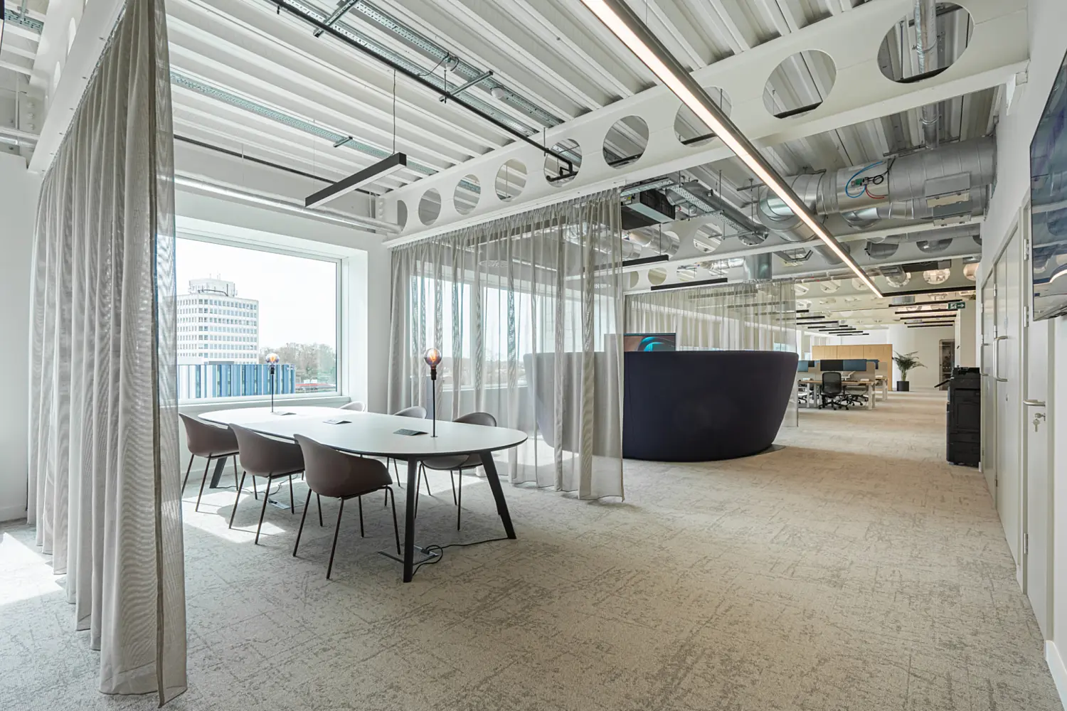 A contemporary open plan office with a round table and four chairs near large windows that fill the space with natural light. Exposed ductwork and lighting fixtures line the ceiling, while semi transparent curtains and a curved partition wall subtly divide zones. The layout is spacious and modern.