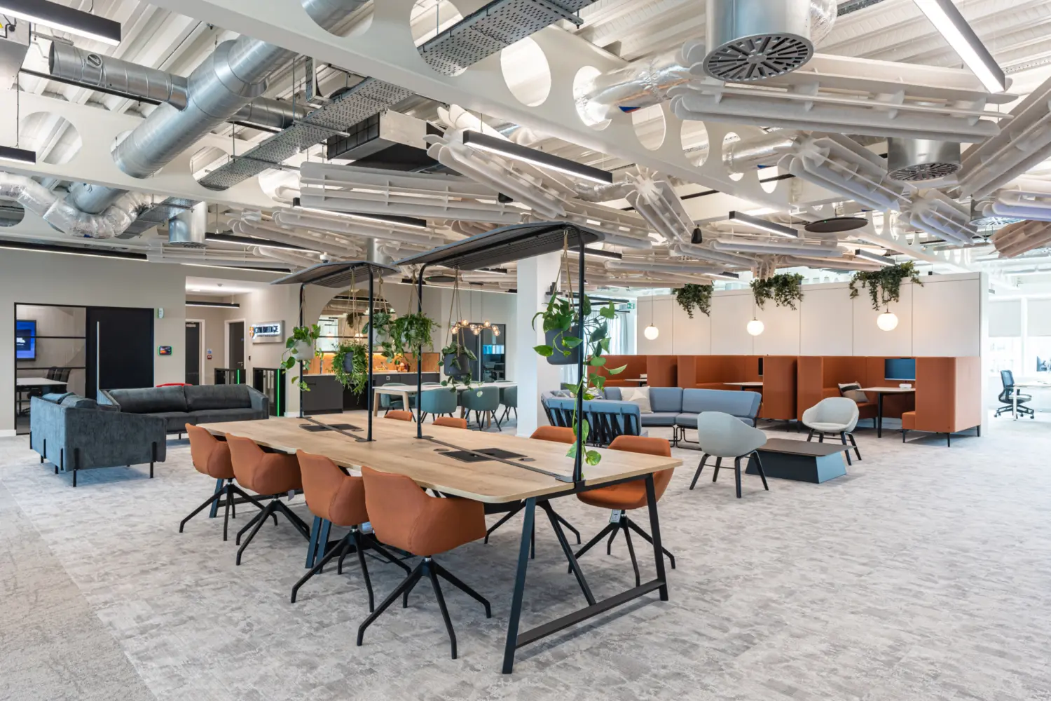 A modern open-plan office with a long wooden table and orange chairs in the foreground, surrounded by hanging plants. Sofas and individual chairs create varied seating zones in the background. Exposed ductwork and lighting fixtures on the ceiling contribute to the contemporary, functional design.