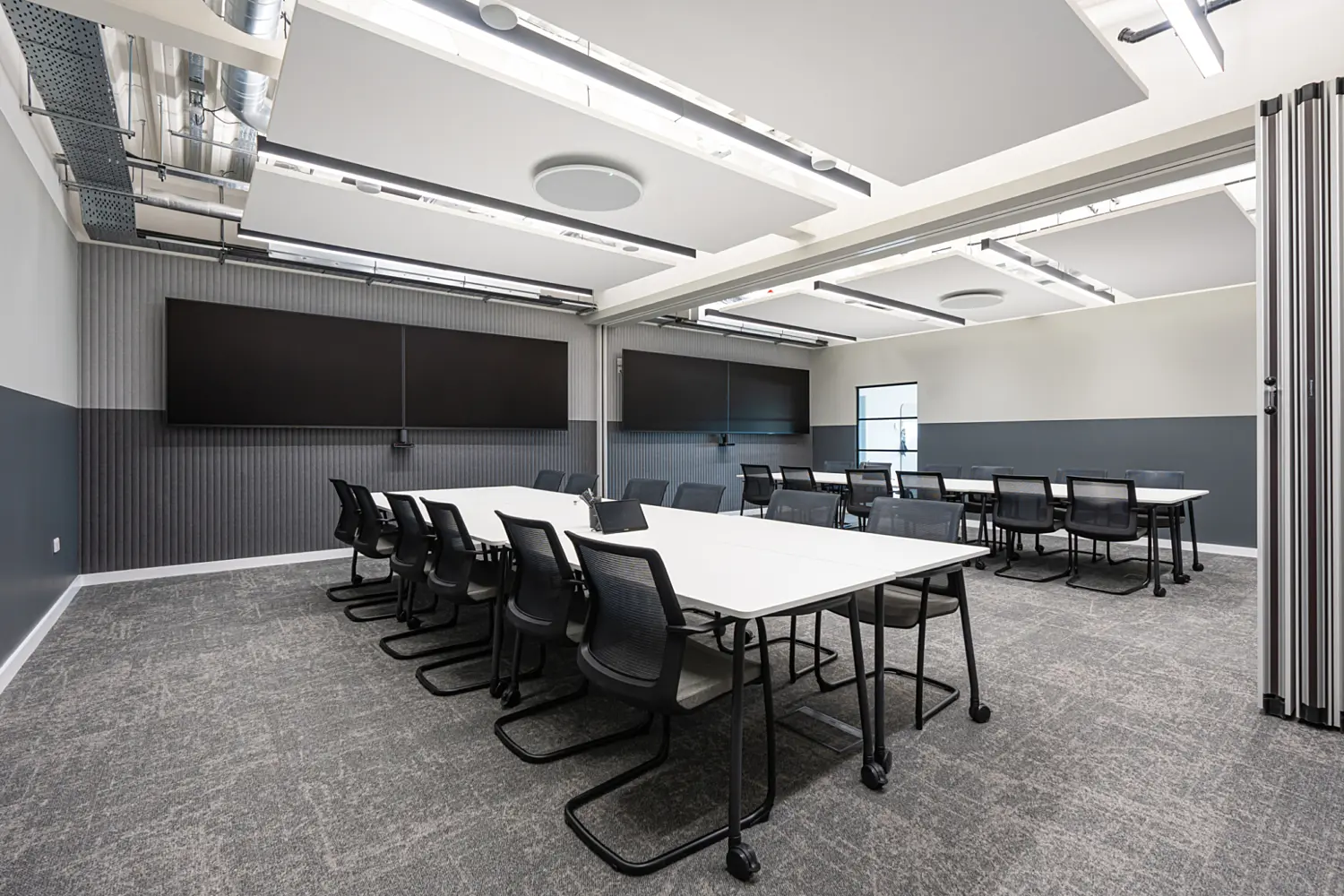 A modern conference room with two large rectangular tables, surrounded by black chairs. The space features a grey and white palette with carpeted flooring, recessed lighting and exposed ductwork. Two wall mounted screens and a movable partition wall add flexibility to the industrial style design.