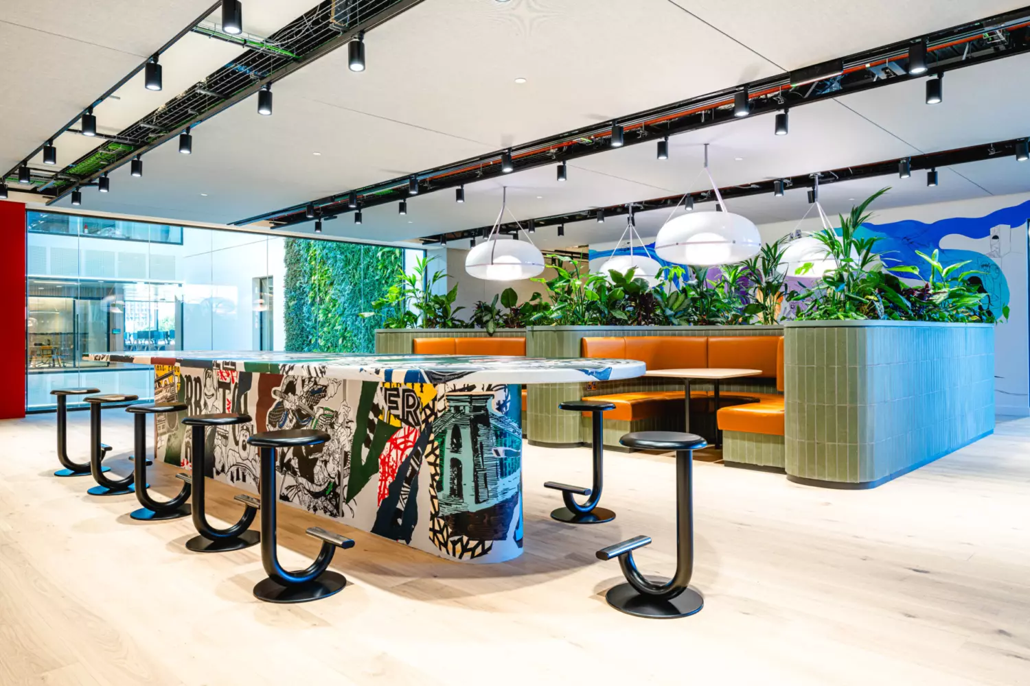 The image shows a modern office fit out and office design in an open-concept communal space. At the centre is a large island table with graffiti-style artwork, surrounded by black stools with U-shaped bases. In the background are green plants and orange cushioned booth seating areas. The ceiling features track lighting and exposed wiring, and a large window lets in natural light.