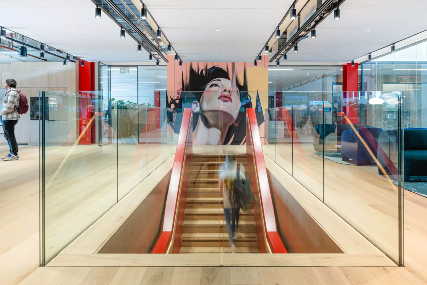 The image shows a modern office fit out and office design. It features an open-plan layout with wooden flooring and glass railings. A staircase with red handrails leads to a lower level, bordered by glass panels. At the top of the stairs is a large mural of a woman's face, adding an artistic element to the space. The ceiling has track lighting and there are several seating areas with contemporary furniture in the background. The overall design highlights openness, light and modern aesthetics.