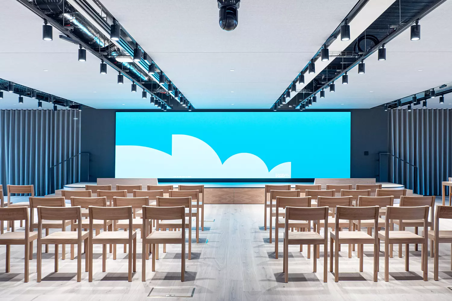 The image shows a modern office fit out and office design in an auditorium space. It features rows of wooden chairs facing a large screen at the front, which displays a blue background with white cloud-like shapes. The space has a minimalist design with light-coloured flooring, ceiling-mounted lights and curtains along the sides.