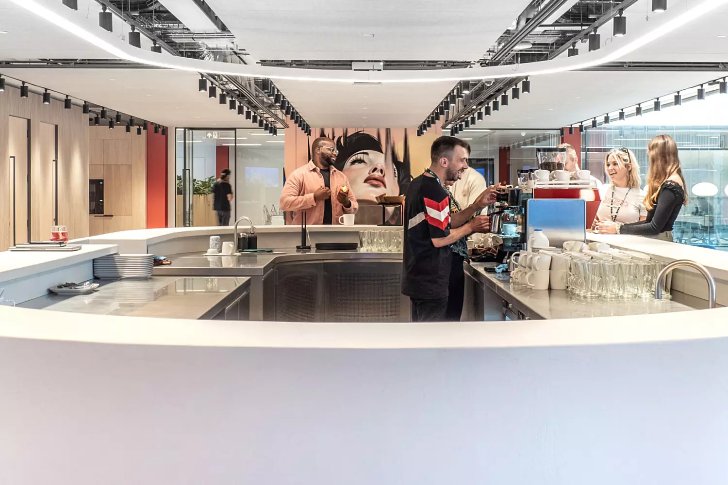 The image shows a modern office fit out and office design in an open-concept coffee bar. It features a curved counter where a barista is preparing drinks and people are gathered, waiting and chatting. The background includes contemporary decor with red accents and glass partitions. The space is brightly lit with track lighting.