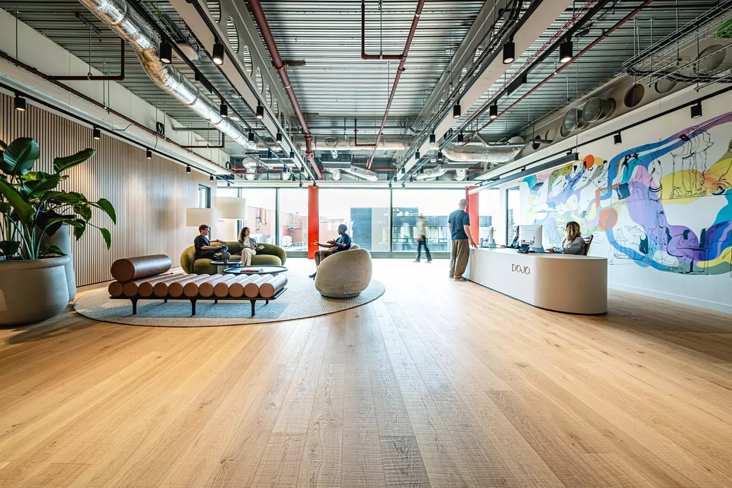 The image shows a modern office fit out and office design in a lobby area. It features a wooden floor and an industrial-style ceiling with exposed ducts and pipes. On the left is a seating area with a round rug, green couches and cylindrical cushions, alongside a large potted plant. On the right is a reception desk with the word "Dojo" on it, where two people are seated. The back wall displays a colourful mural and large windows and glass doors lead to another part of the building.