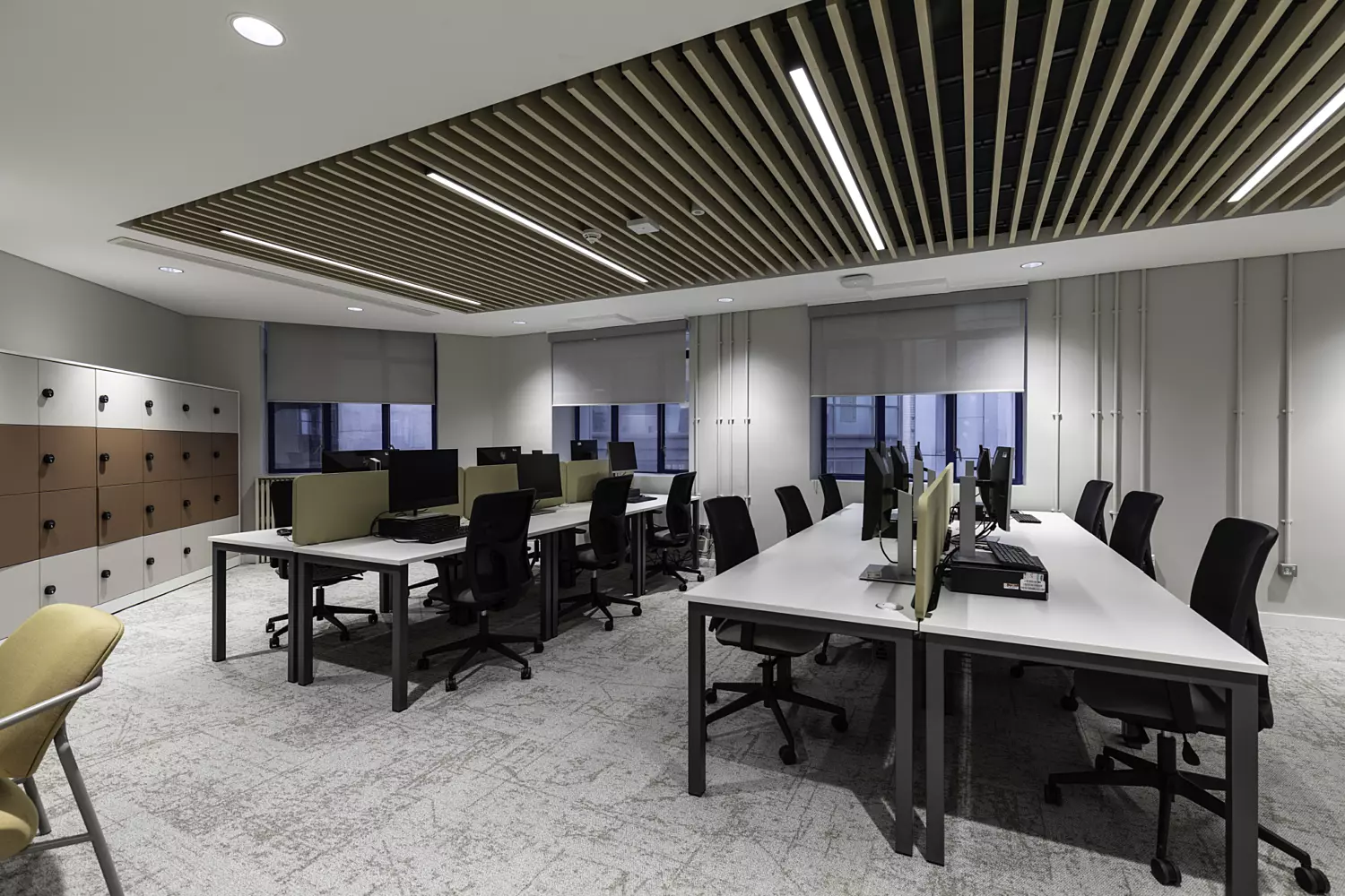 Modern office with rows of desks, computer monitors, slatted wooden ceiling, and lockers along the wall.