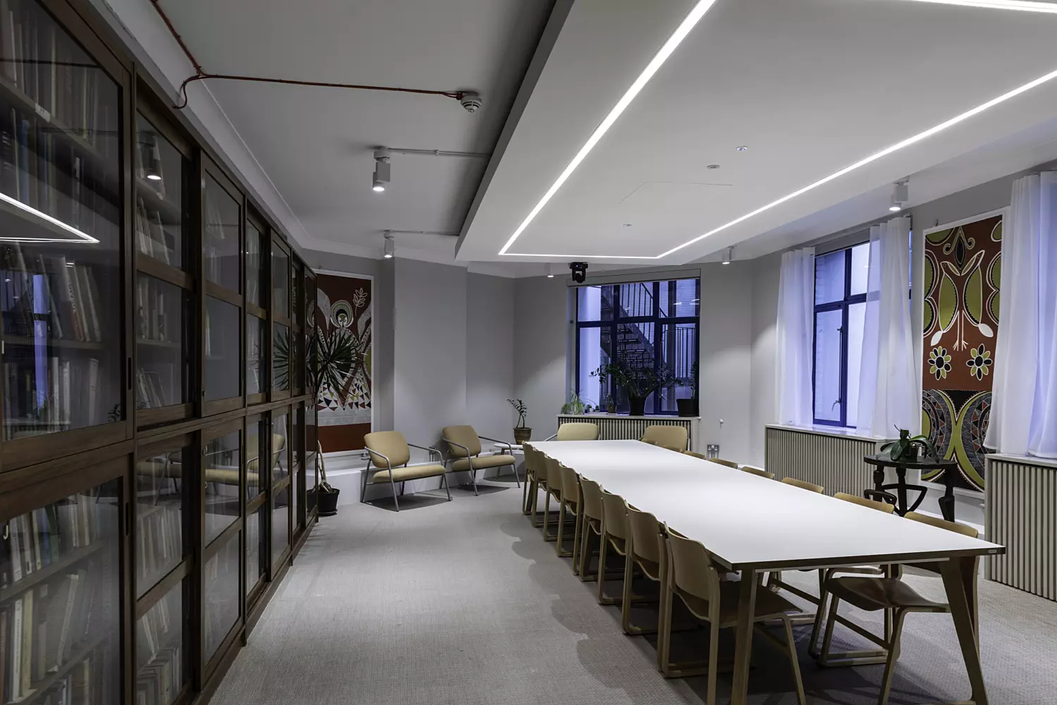 Conference room with a long white table, wooden chairs, bookshelves, and large windows with sheer curtains.