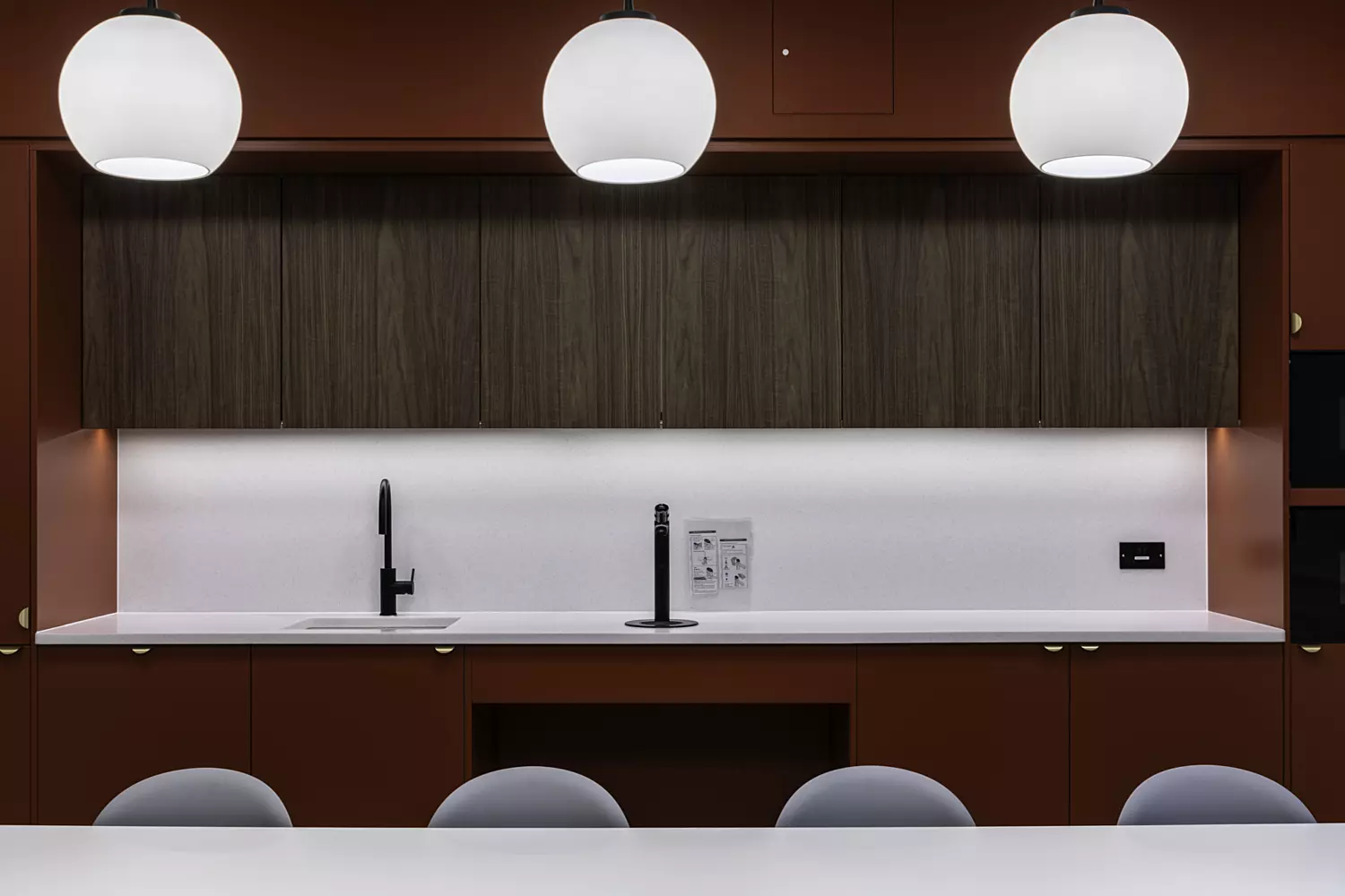 A modern kitchen interior featuring red cabinets and three large, round pendant lights hanging from the ceiling. The countertop is white with a black faucet and sink on the left side. Above the countertop are wooden cabinets, and below it are more red cabinets with gold handles. There is a small black appliance on the right side of the counter, possibly an oven or microwave. Four grey chairs are partially visible at the bottom of the image, suggesting a dining area in front of the kitchen counter.