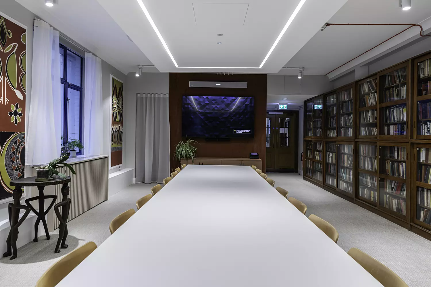 Contemporary meeting room with a long white table, brown chairs, wall-mounted TV, and bookshelves.