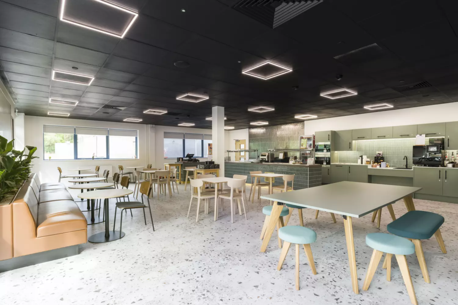 A bright, modern office cafeteria featuring mixed seating options including tables with chairs, a long wall bench and a communal table with stools. Square ceiling lights and large windows provide ample illumination. At the back, a counter area houses coffee machines and kitchen equipment, blending functionality with a welcoming atmosphere.