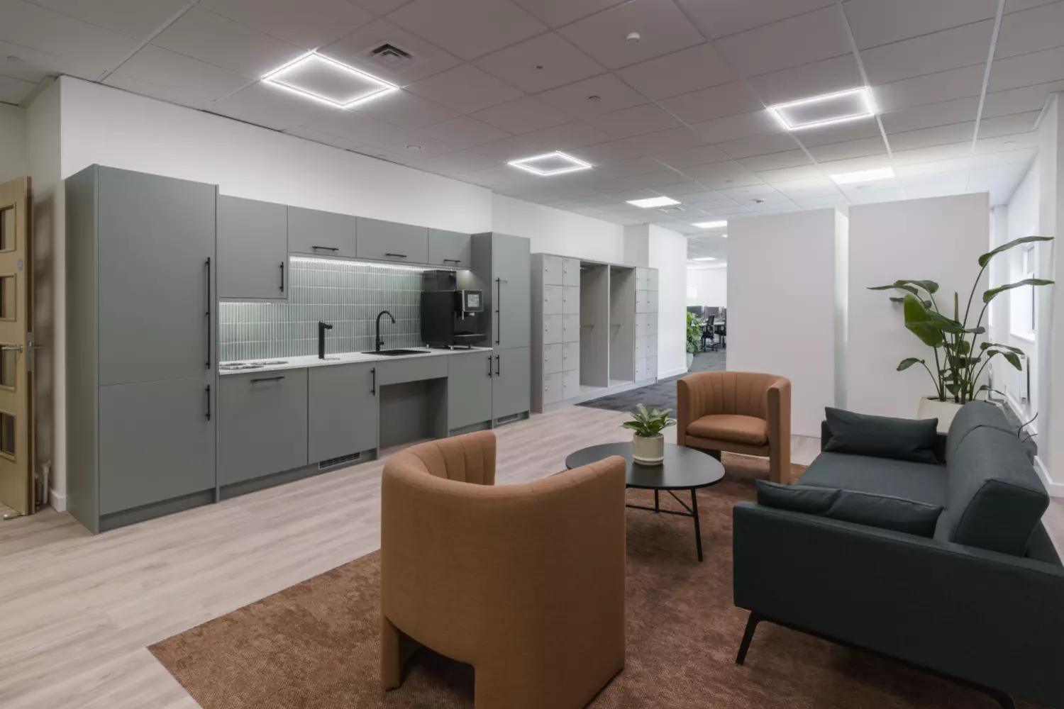 A modern office lounge with a green kitchenette featuring a sink and coffee machine. The seating area includes two brown armchairs, a green sofa and a round black coffee table on a brown rug. Light wood and grey carpet flooring define zones, while square LED ceiling lights and a potted plant add warmth.