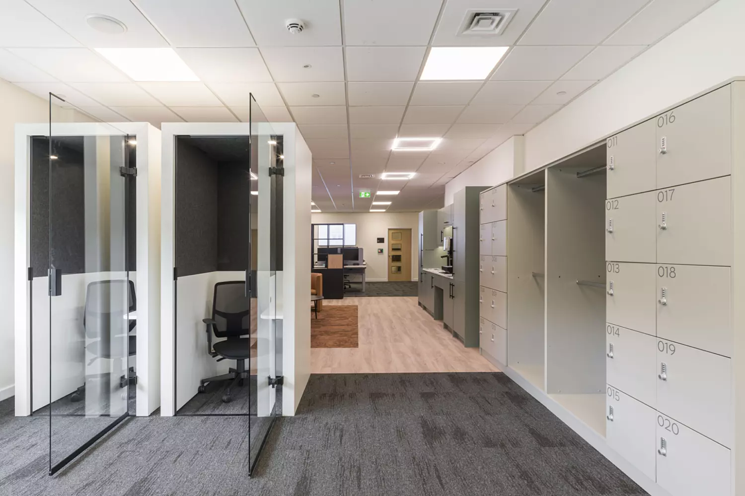 A modern office corridor featuring two glass-walled cubicles with office chairs on the left and numbered lockers on the right. The flooring transitions from carpet to wood, adding visual interest. Recessed ceiling lights provide clean illumination, while the hallway opens into a larger workspace with visible office furniture.