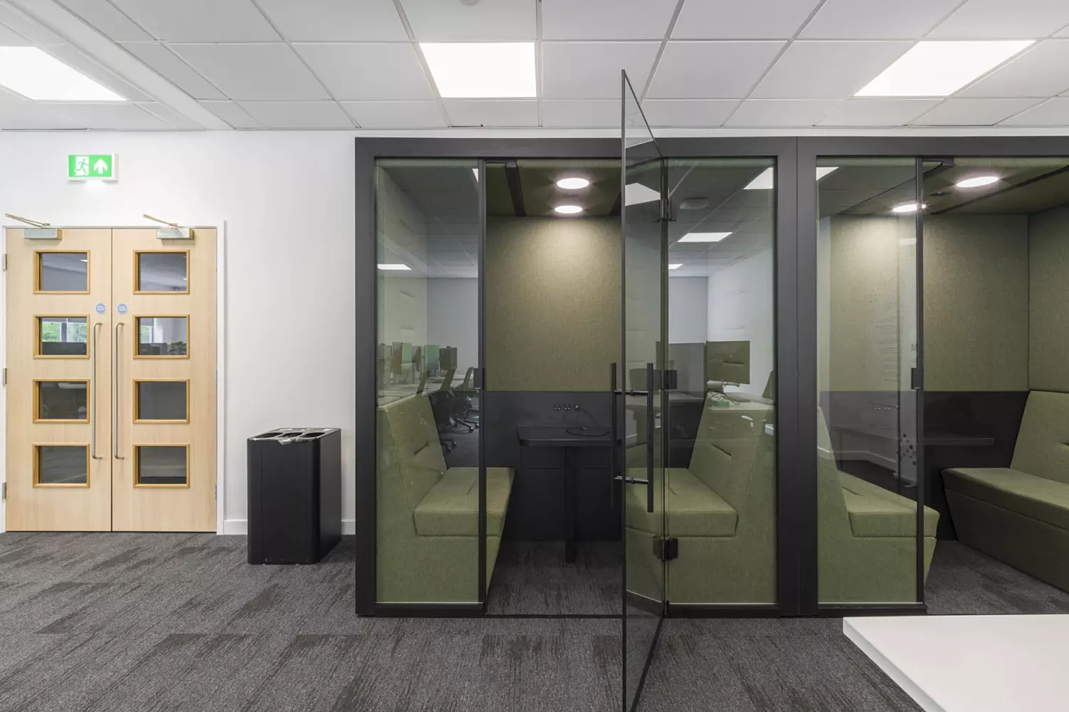 A modern office space featuring two glass-enclosed meeting pods with green cushioned seating and small tables. One pod has its door open, the other closed. Overhead ceiling lights illuminate the area, which has grey patterned carpet. To the left, wooden double doors with an emergency exit sign add functional contrast.