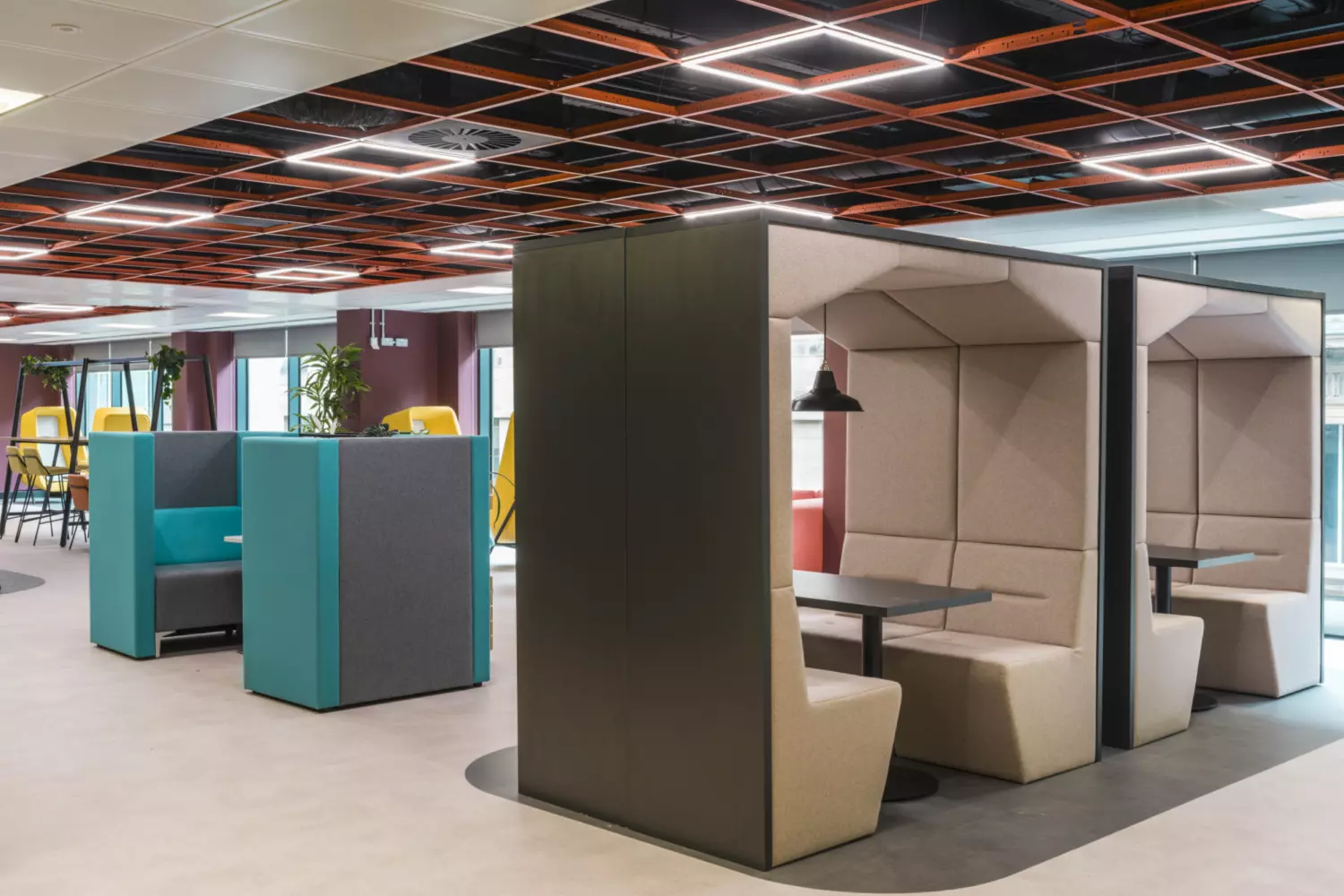 The image shows a modern office fit out and office design with a variety of seating arrangements. Two enclosed booth-style seating areas with cushioned benches and tables are positioned for private meetings or focused work. The booths have a beige colour scheme and overhead lighting. In the background are teal and grey high-backed chairs and yellow chairs around tables. The ceiling features an orange grid pattern with integrated lighting and large windows on the right bring in natural light.