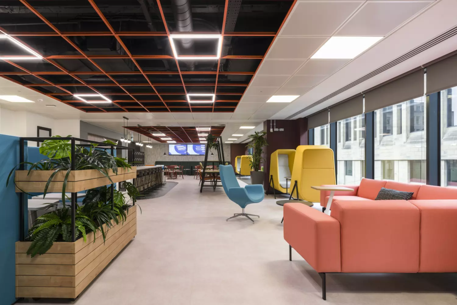 The image shows a modern office fit out and office design with a variety of seating options and decor. On the left are plants in wooden planters, a blue chair sits in the centre and a pink couch is on the right. In the background are yellow and green enclosed seating areas, tables, chairs and large windows that let in natural light. The ceiling features an orange grid pattern with recessed lighting.