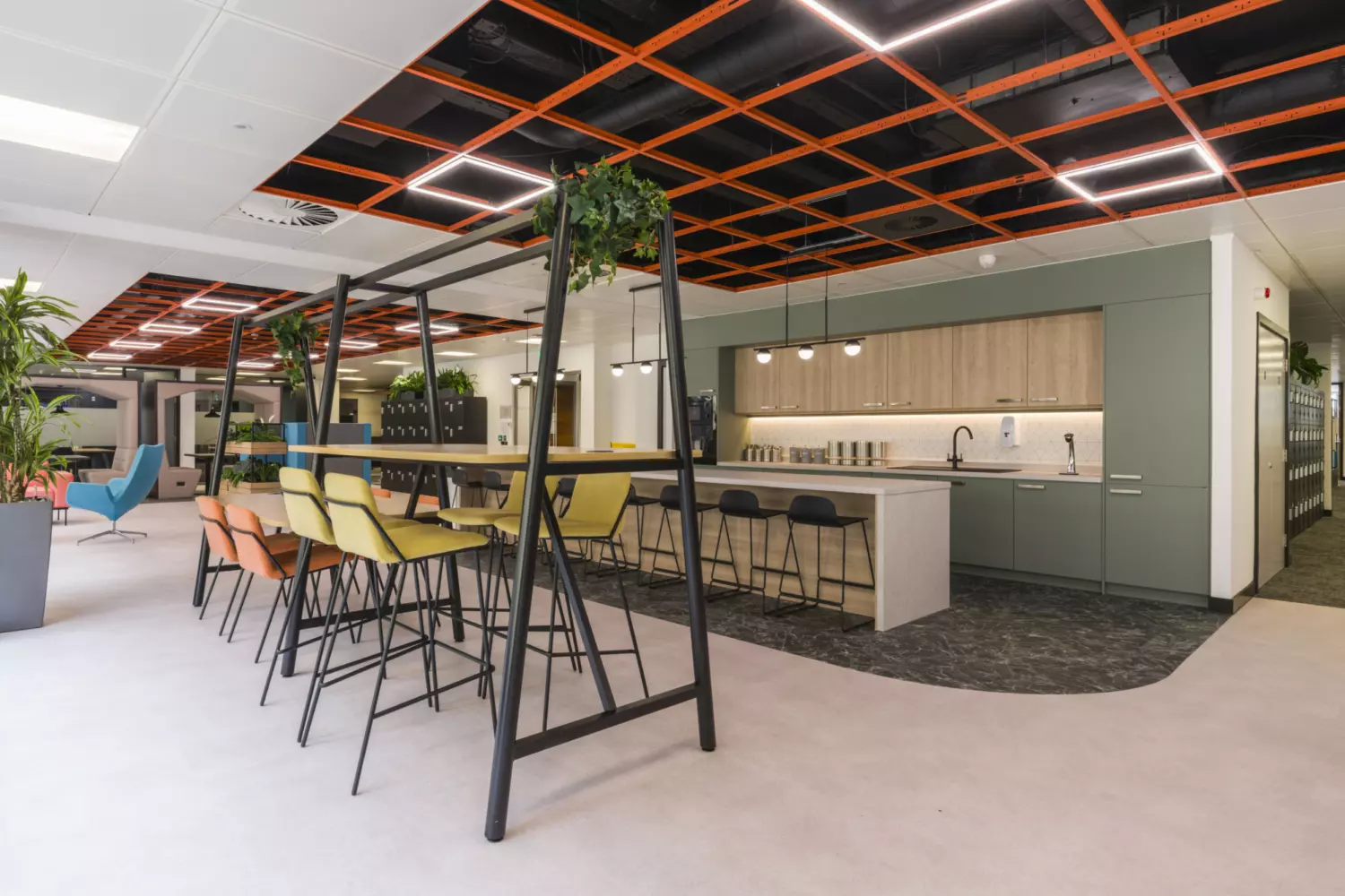 The image shows a modern office fit out and office design in a kitchen and dining area. A long high table runs through the centre, with yellow and orange bar stools on one side and black bar stools on the other. The ceiling has an open design with an orange grid pattern. The kitchen area includes a countertop, sink, coffee machines and hanging lights, with plants placed around the space for decoration.