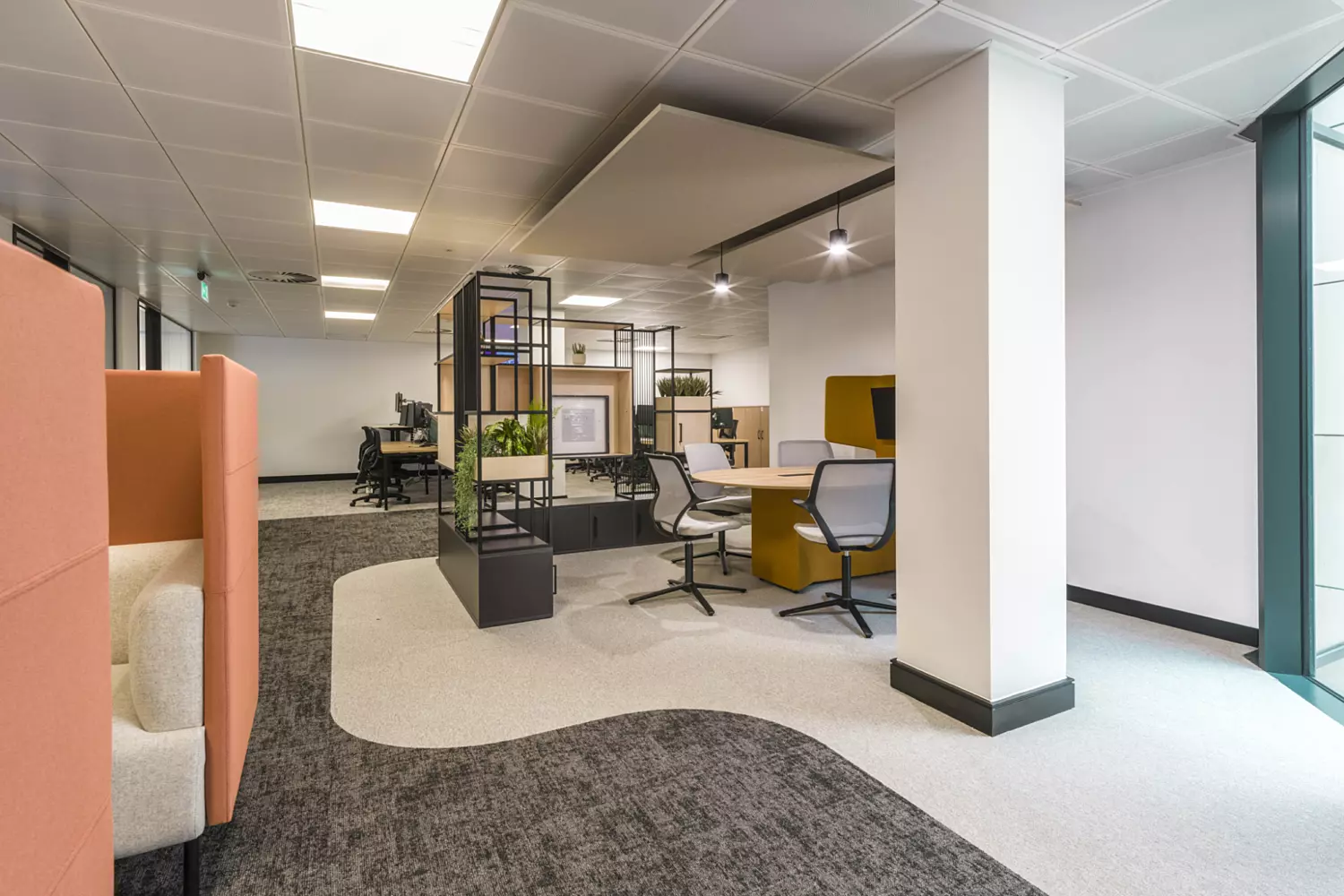 The image shows a modern office fit out and office design in an open-plan workspace. It includes several workstations with chairs, desks and computer monitors. The flooring is carpeted in a mix of colours and patterns. There are partitioned areas with orange and beige seating, and plants are placed on shelves and desks for decoration. The ceiling has recessed lighting and a large window on the right lets in natural light.