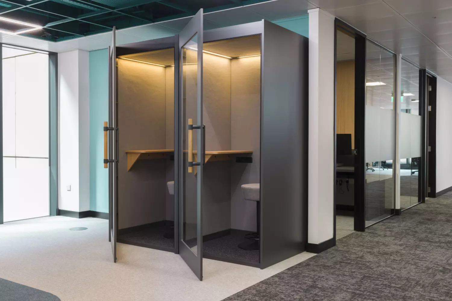 The image shows a modern office fit out and office design featuring two small enclosed work booths. Each booth has a glass door, a wooden desk and an upholstered chair, designed for individual work or private calls. The surrounding area includes glass walls and doors leading to other parts of the office. The floor is carpeted and the ceiling has exposed elements with lighting fixtures.