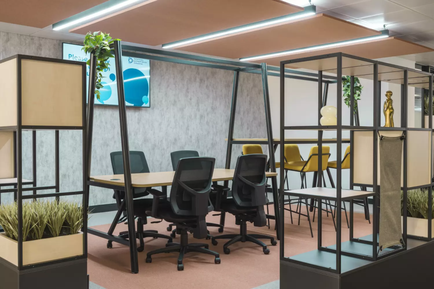 The image shows a modern office fit out and office design featuring a conference area with a table surrounded by six black office chairs. Shelves with decorative items and plants are arranged around the space. In the background is another seating area with yellow chairs and a high table. The ceiling has linear LED lights and a screen on the wall displays graphics.