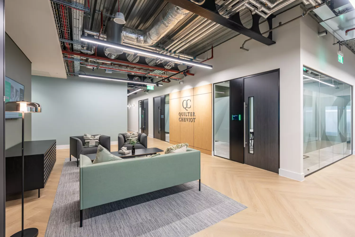 A modern office lobby with a light green sofa, two grey armchairs and a coffee table on a grey rug. The space features light-coloured walls, exposed ceiling ductwork and a mix of glass panelled and wooden doors, including one with the Quilter Cheviot logo, creating a clean, professional welcome area.