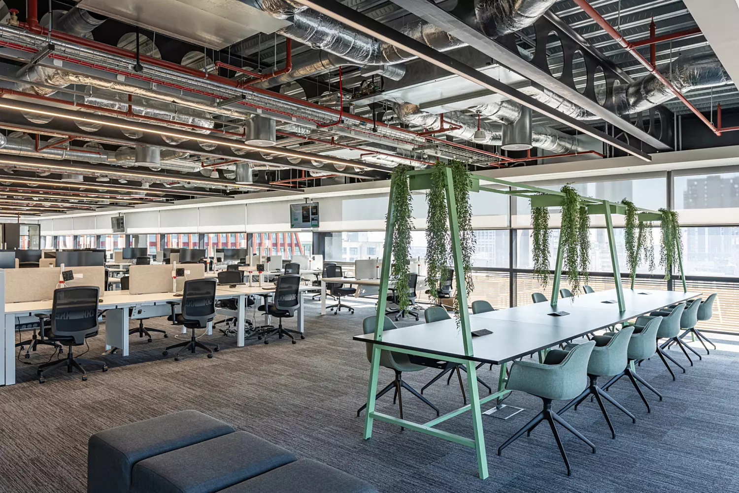 A modern open plan office with rows of workstations featuring desks, office chairs, computer monitors and partitions. The ceiling has exposed ductwork and piping, contributing to an industrial look. In the foreground, a long table with green framework is positioned beneath hanging plants, surrounded by chairs. Large windows allow natural light to brighten the space.