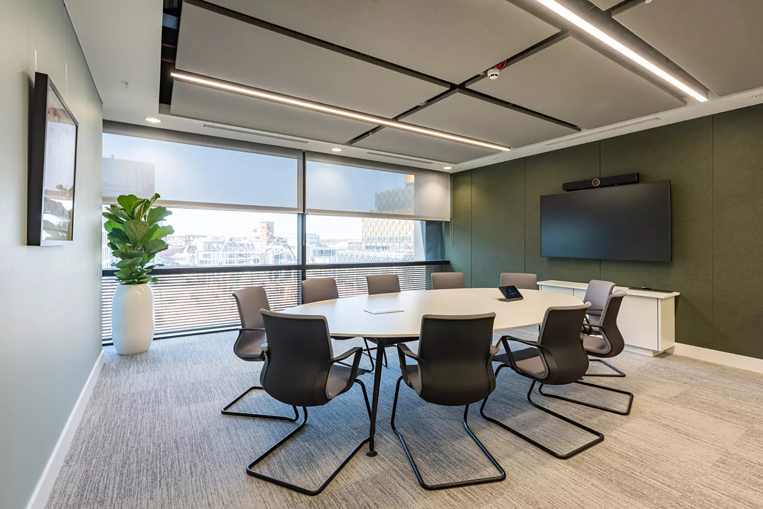 A modern conference room featuring a large oval table surrounded by eight black chairs. A large window with partially drawn blinds lets in natural light. A potted plant sits in the corner near the window. One wall has a mounted flat screen monitor above a white cabinet. The ceiling includes recessed lighting and acoustic panels. The floor is covered in light grey carpet.