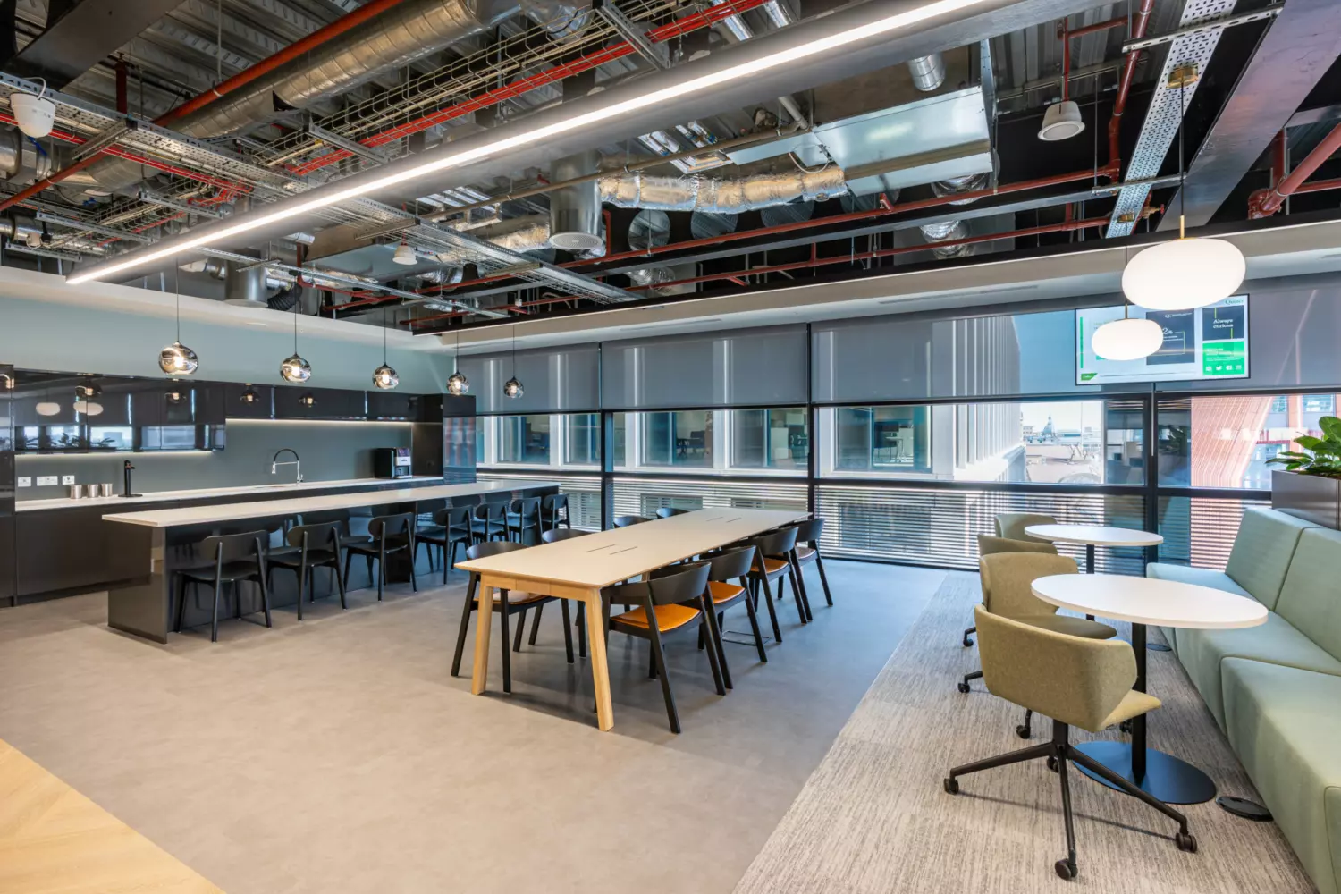 A modern office break room or communal area with an industrial design featuring exposed ductwork and piping on the ceiling. The space includes a long table with chairs, a kitchen area with bar stools and additional seating with small round tables and chairs. Large windows allow natural light to fill the room.