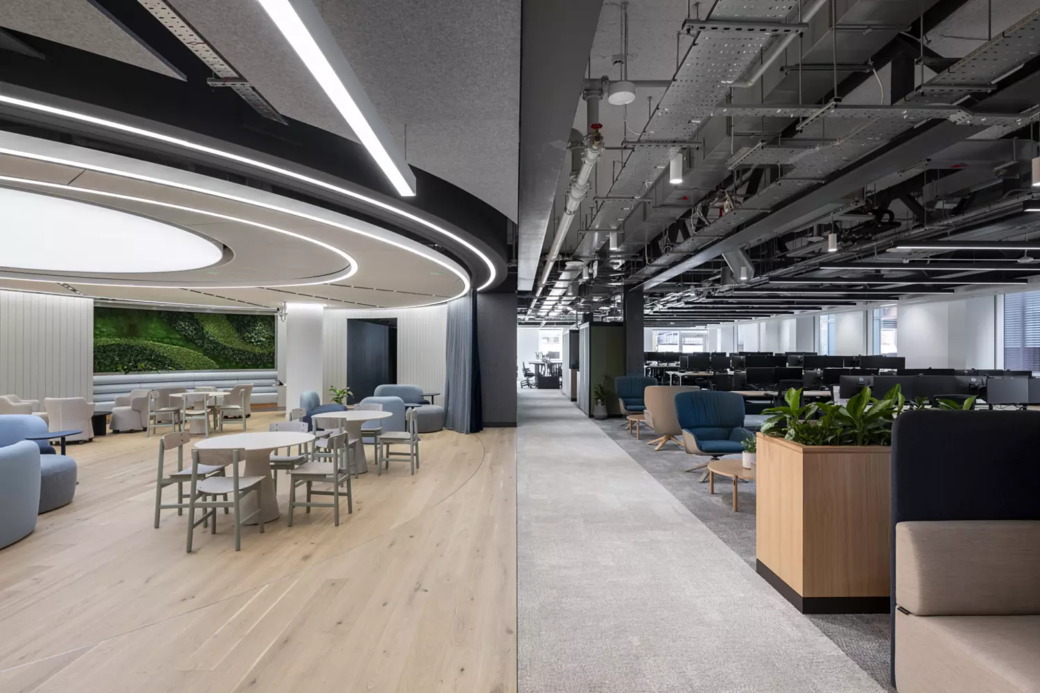 A modern office interior split into two distinct areas. On the left side, there's a bright, stylish lounge space with light wood flooring, soft pastel chairs, round tables, and a green living wall in the background. The ceiling features circular recessed lighting that adds a sleek, futuristic feel. On the right side, there's an open-plan workspace with grey carpet, rows of black computer monitors on desks, and exposed industrial ceiling elements like pipes and cables. This area also includes some casual seating with blue and beige armchairs, coffee tables and planter boxes with greenery.