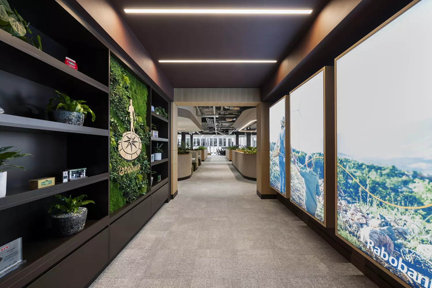 Modern office hallway with a carpeted floor and ceiling lights. On the left, shelves hold plants and decorative items, including a large green wall with the Rabobank logo. On the right, illuminated panels display nature scenes and the Rabobank name. The hallway leads to an open office space in the background.