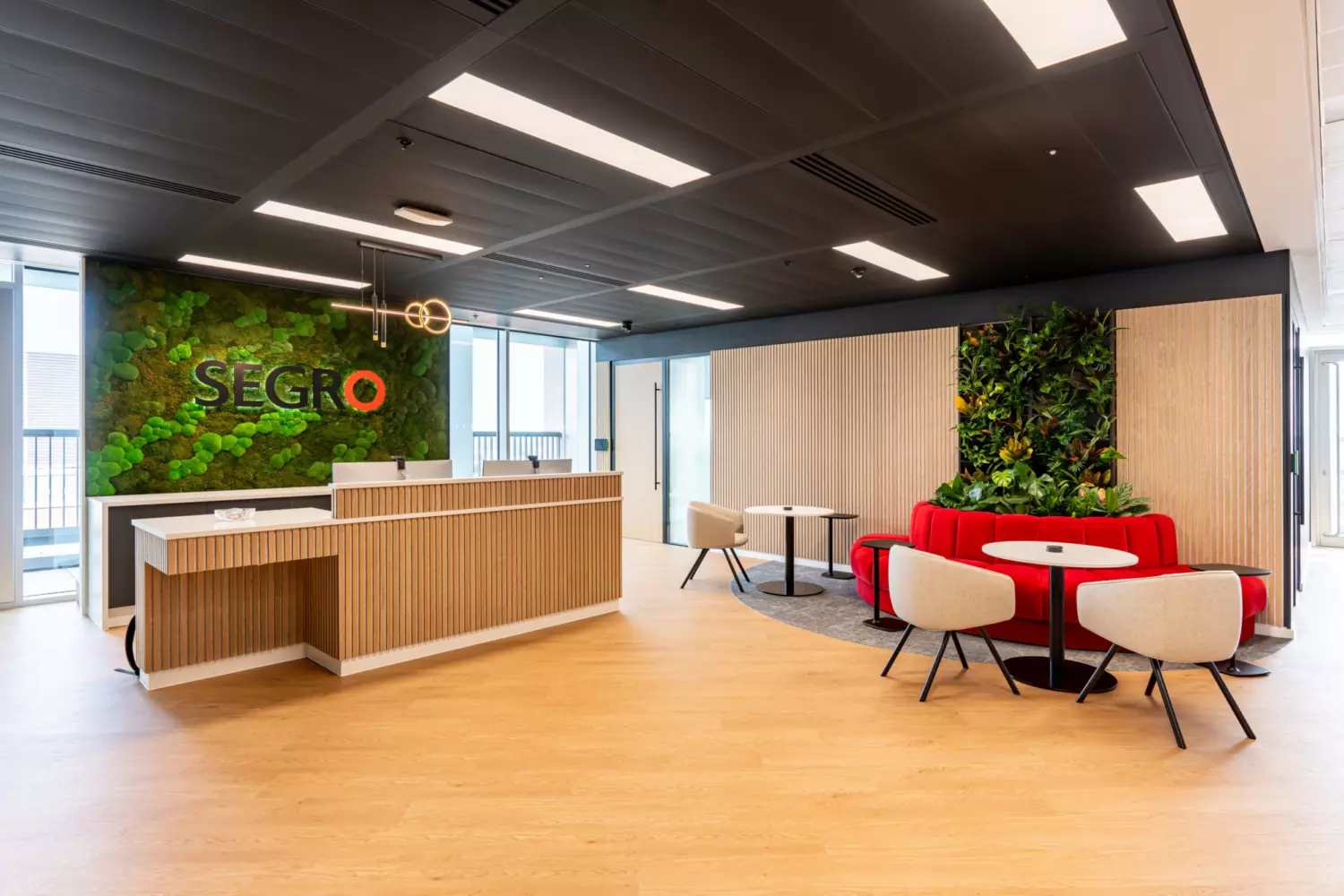 A modern office reception area with wooden flooring and ceiling. On the left, a wooden reception desk with white accents sits in front of a green wall displaying the text "SEGRO". On the right, a seating area includes red cushioned seats around small round tables with beige chairs. Another green wall with plants is positioned behind the seating area. Large windows allow natural light to enter the space.
