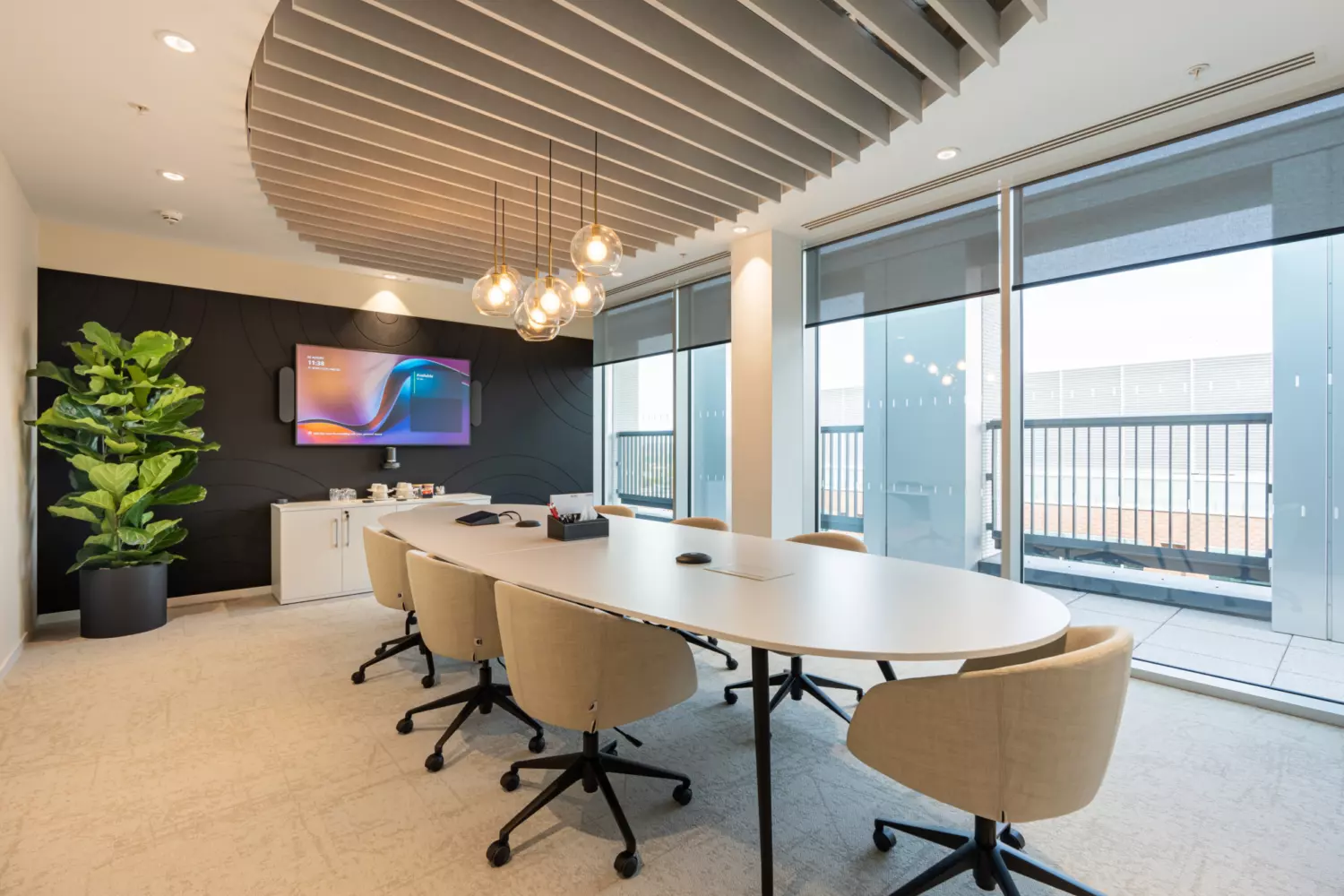 A modern meeting room with a long oval table surrounded by eight beige wheeled office chairs. A large flat-screen TV is mounted on a black accent wall at the far end, displaying colourful graphics. Below it, a white cabinet holds various items and a large green plant in a black pot sits to the left. The ceiling features an artistic arrangement of hanging light fixtures with multiple round bulbs. Floor-to-ceiling windows on one side let in natural light and offer views of an outdoor area with railings and buildings.