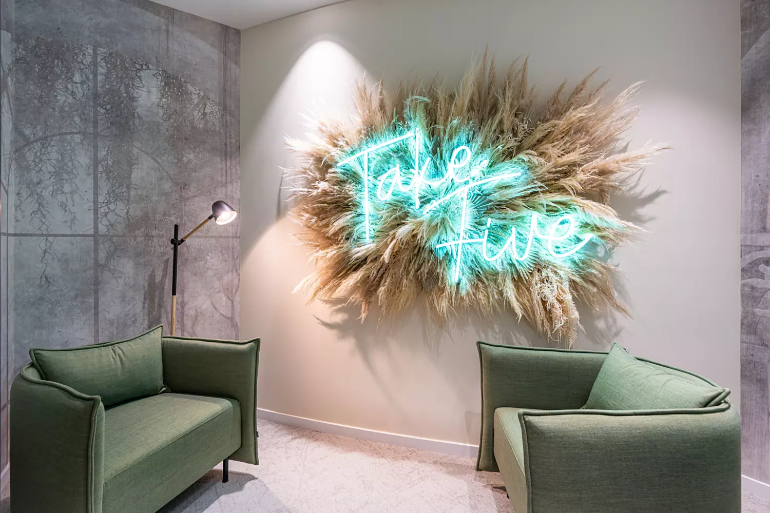 A modern cosy seating area featuring two green armchairs facing each other. Between them is a wall with a decorative display of dried plants and a neon sign that reads "Take Five". A floor lamp on the left casts light onto the arrangement, adding warmth to the space.