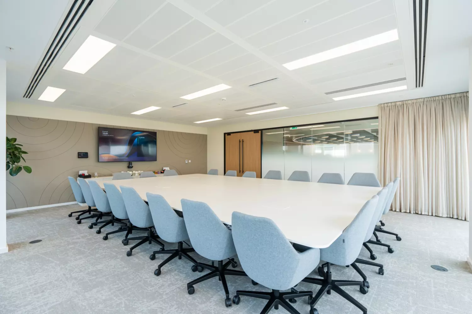 modern conference room with a large rectangular table surrounded by light blue wheeled office chairs. The space features a minimalist design with white walls and ceiling, recessed lighting and a wall-mounted flat-screen TV. A plant sits in the corner and glass doors with curtains line one side of the room.