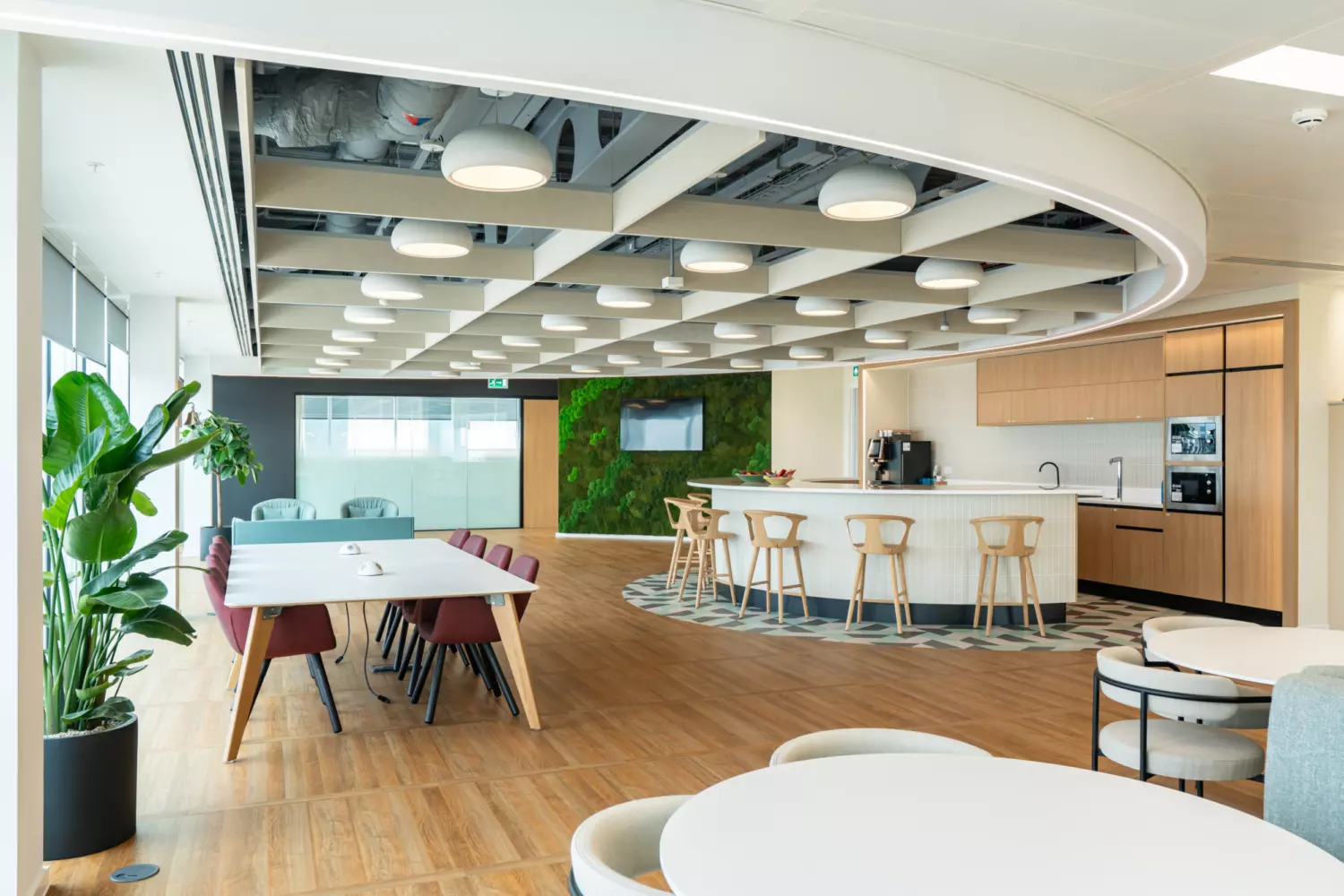 A modern open-concept office casual seating area with wooden flooring and a mix of seating including high stools at a counter and lower chairs around dining tables. A large plant is visible in the foreground and a green wall with plants forms part of the background. The ceiling features exposed beams and circular light fixtures. A kitchen area with cabinets, appliances and a countertop is also present for food or drink preparation.