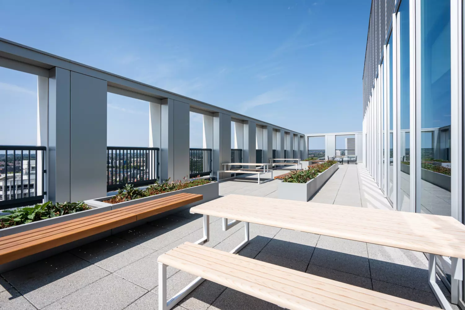 A rooftop terrace with a modern architectural design featuring long wooden benches and picnic tables arranged along the space. Planters filled with greenery line the terrace. Large rectangular openings in the wall offer views of the cityscape. The sky is clear and blue.