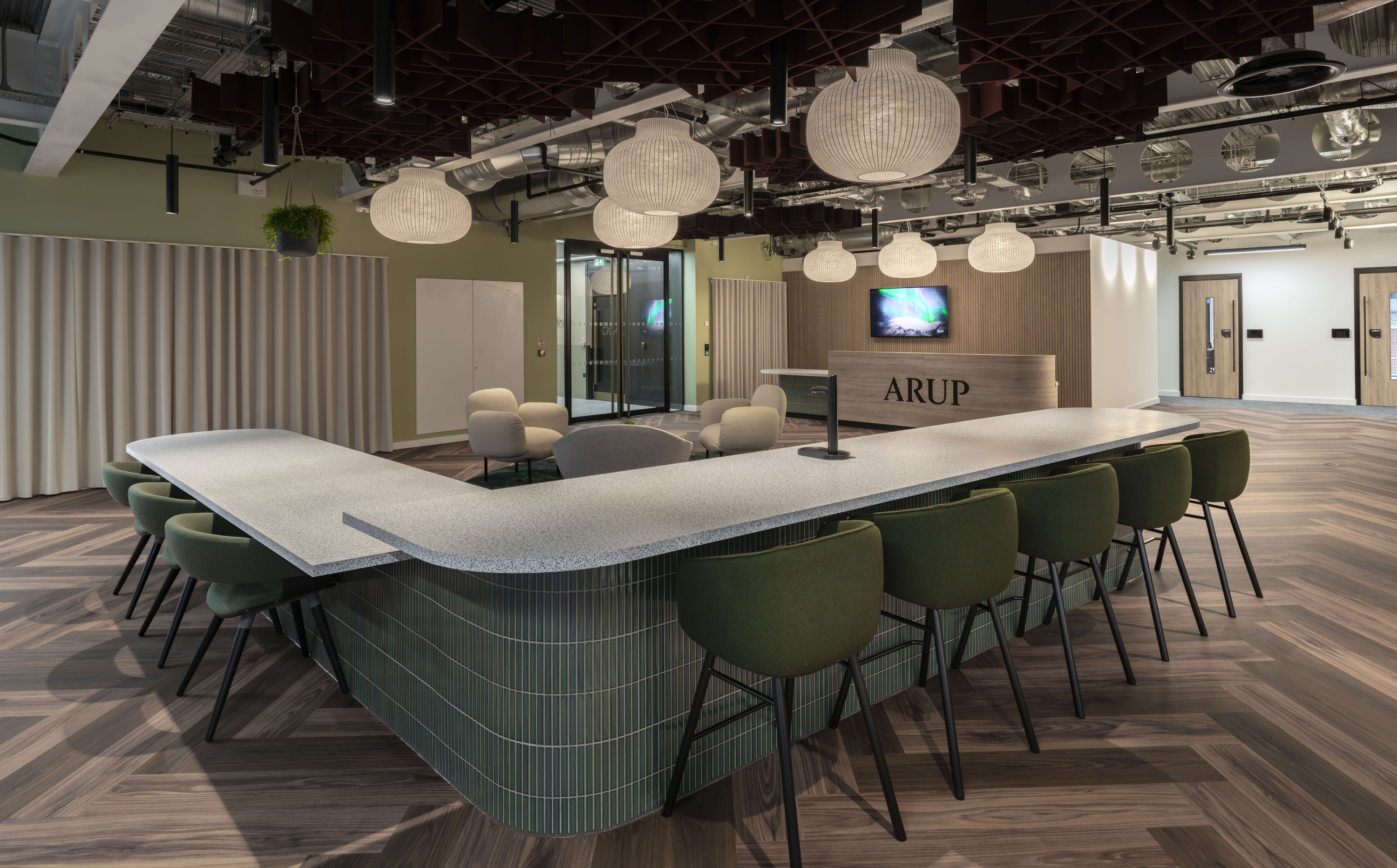 A contemporary office interior featuring a large, curved communal table with a sleek white surface, surrounded by green upholstered chairs. Overhead, spherical pendant lights hang from a ceiling with exposed industrial-style piping and ductwork. The floor is laid with herringbone-patterned wooden flooring. In the background, a glass-walled meeting room and a reception area are visible, with the word "ARUP" displayed on the front desk. The walls are painted in neutral tones, with vertical wooden slats adding texture and warmth to one section.
