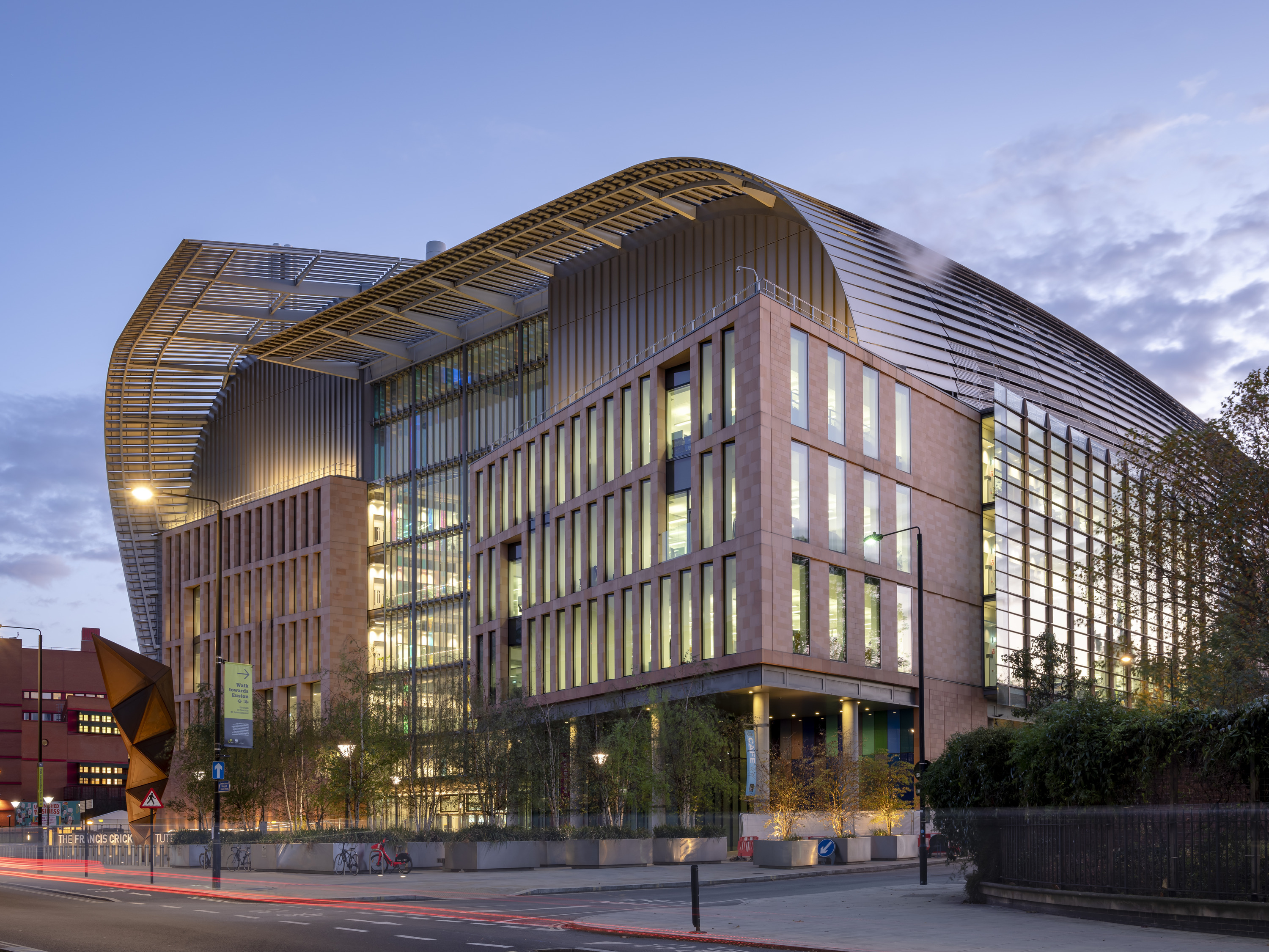Architecturally distinctive building with a curved roof and large glass windows spanning multiple storeys. The exterior combines glass and brick materials, creating a visually striking contrast. In the foreground, trees and streetlights line the street, adding context to the urban setting