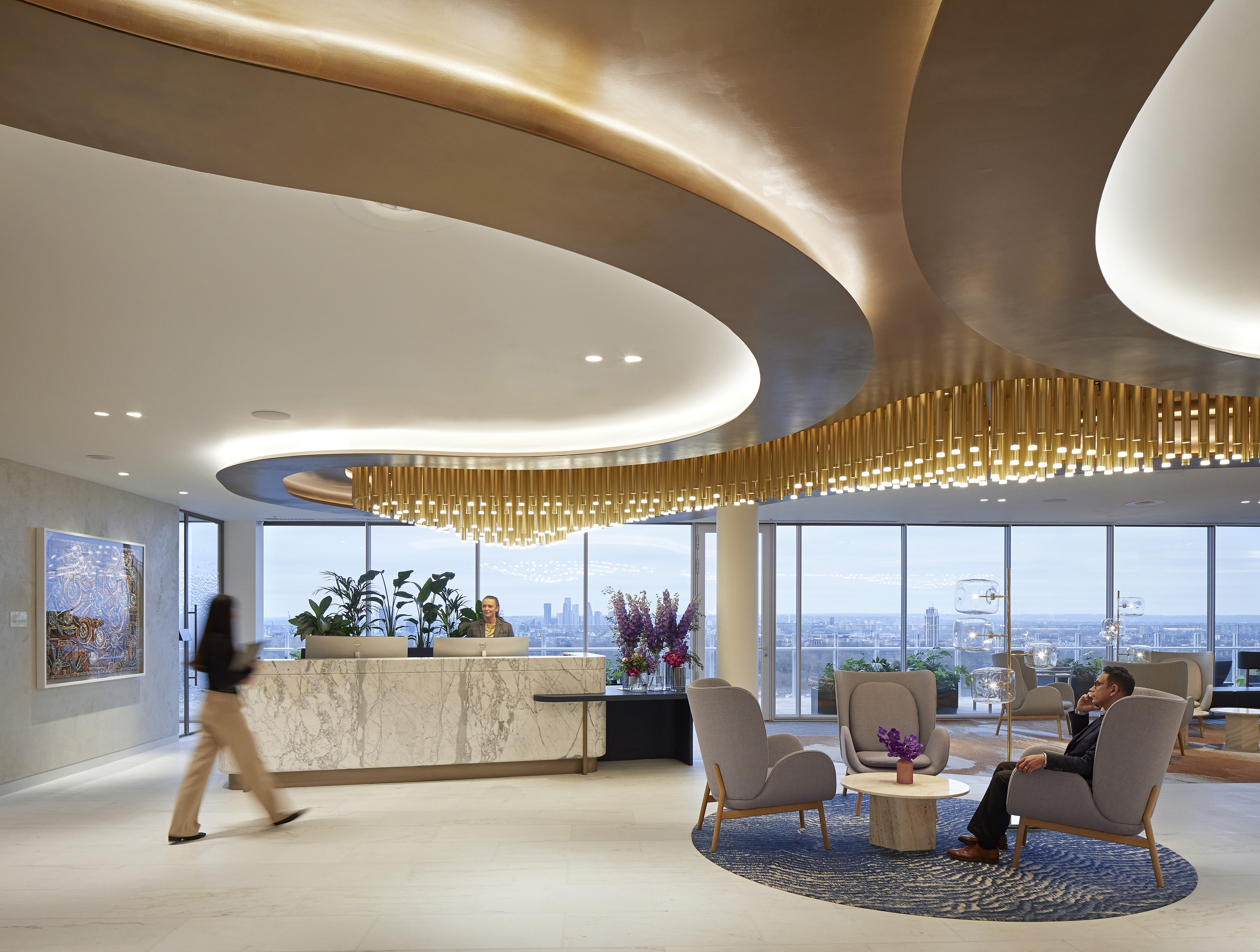 Modern office lobby with a refined and elegant design, featuring a reception desk on the left and a large curved ceiling installation adorned with hanging golden elements. The space includes a seating area with two chairs and a small table topped with flowers, creating a welcoming atmosphere. Large windows at the back offer views of an urban skyline, while the overall design blends luxury with contemporary minimalism.