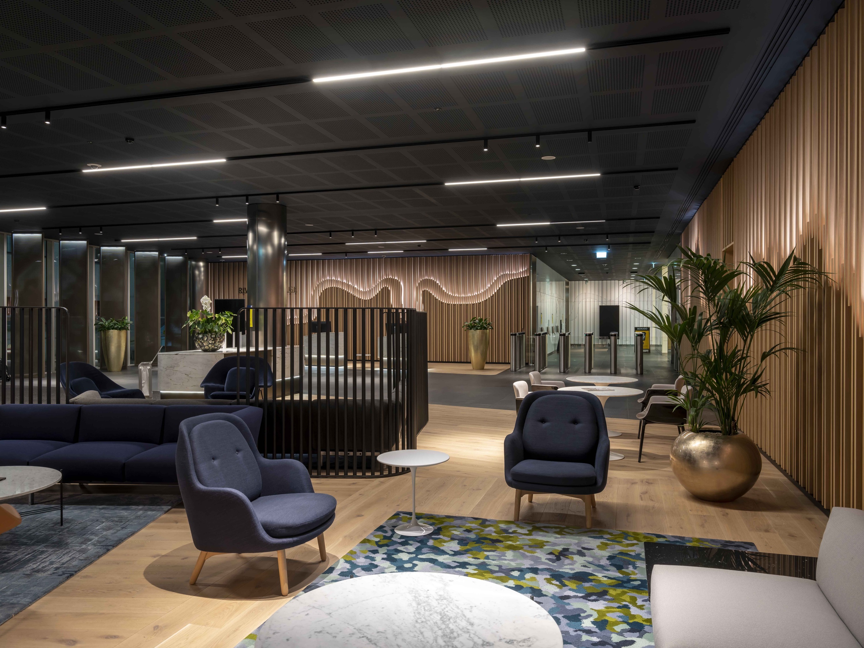 A modern, stylish interior space featuring contemporary furniture including blue armchairs and sofas. A patterned rug and a large potted plant are in the foreground. The background includes wooden wall panelling, ceiling lights and a partition with vertical slats dividing the room into separate areas.