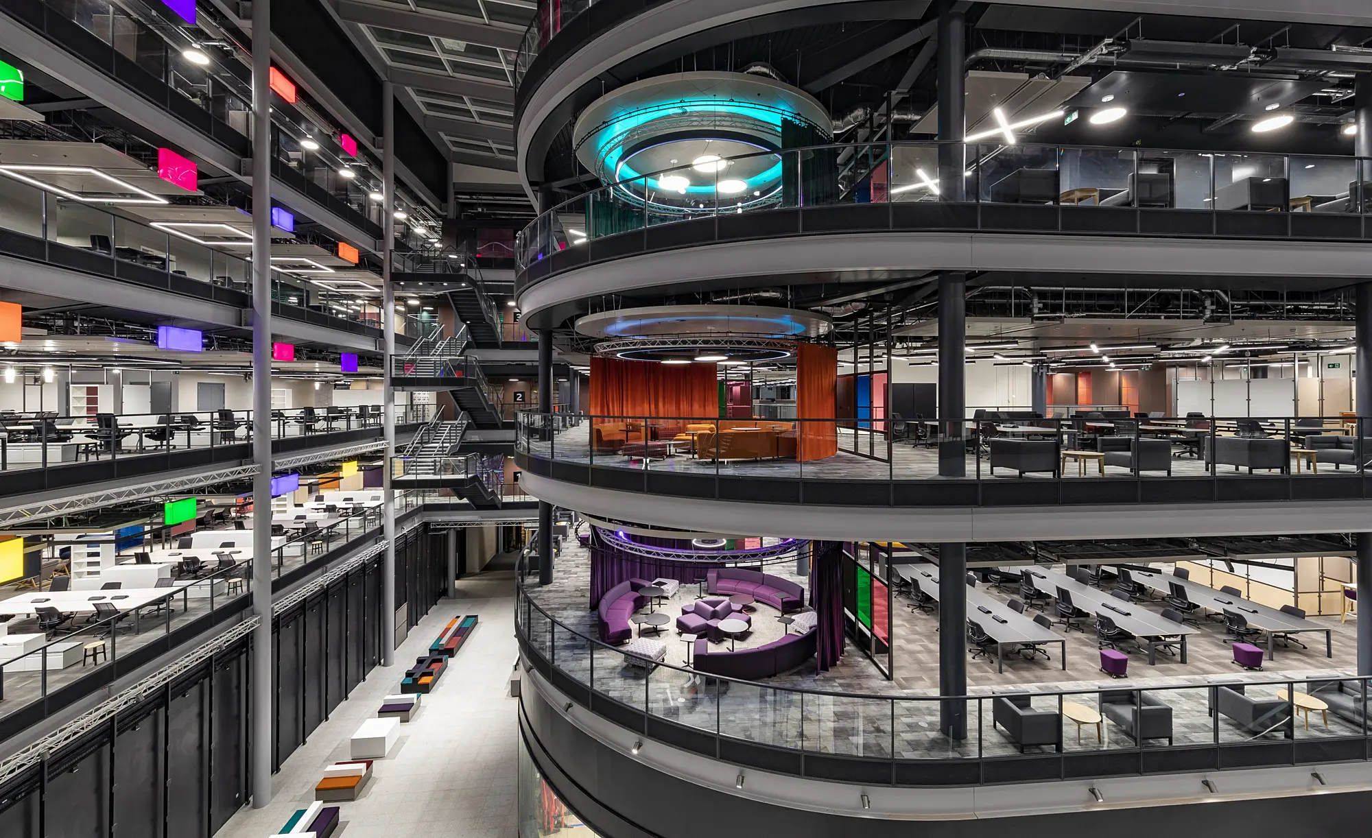 The interior of a modern, multi level office building with open plan workspaces. Several floors feature circular balconies overlooking a central atrium. Each level includes seating areas, desks and meeting spaces. The space is brightly lit with colourful accents throughout.