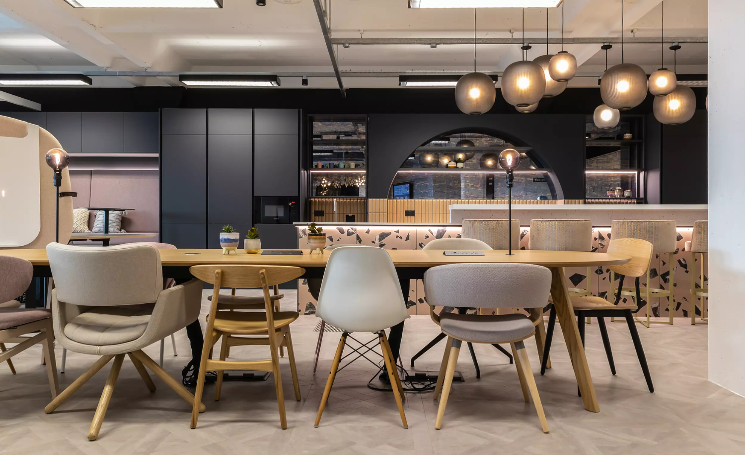 An office's modern dining area featuring a long wooden table surrounded by assorted chairs. In the background, there is a kitchen with dark cabinets, open shelves and appliances. Several spherical pendant lights hang from the ceiling above the table.