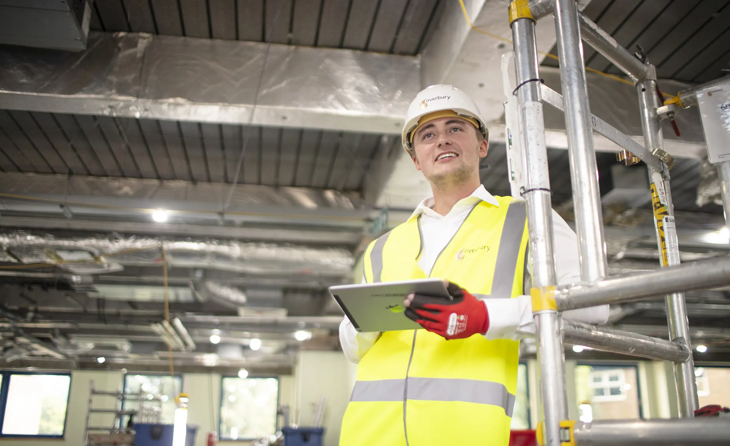 Person in a high visibility yellow vest and white hard hat stands beside metal scaffolding in a construction site, holding a tablet with red work gloves, with exposed ductwork and ceiling services visible overhead.
