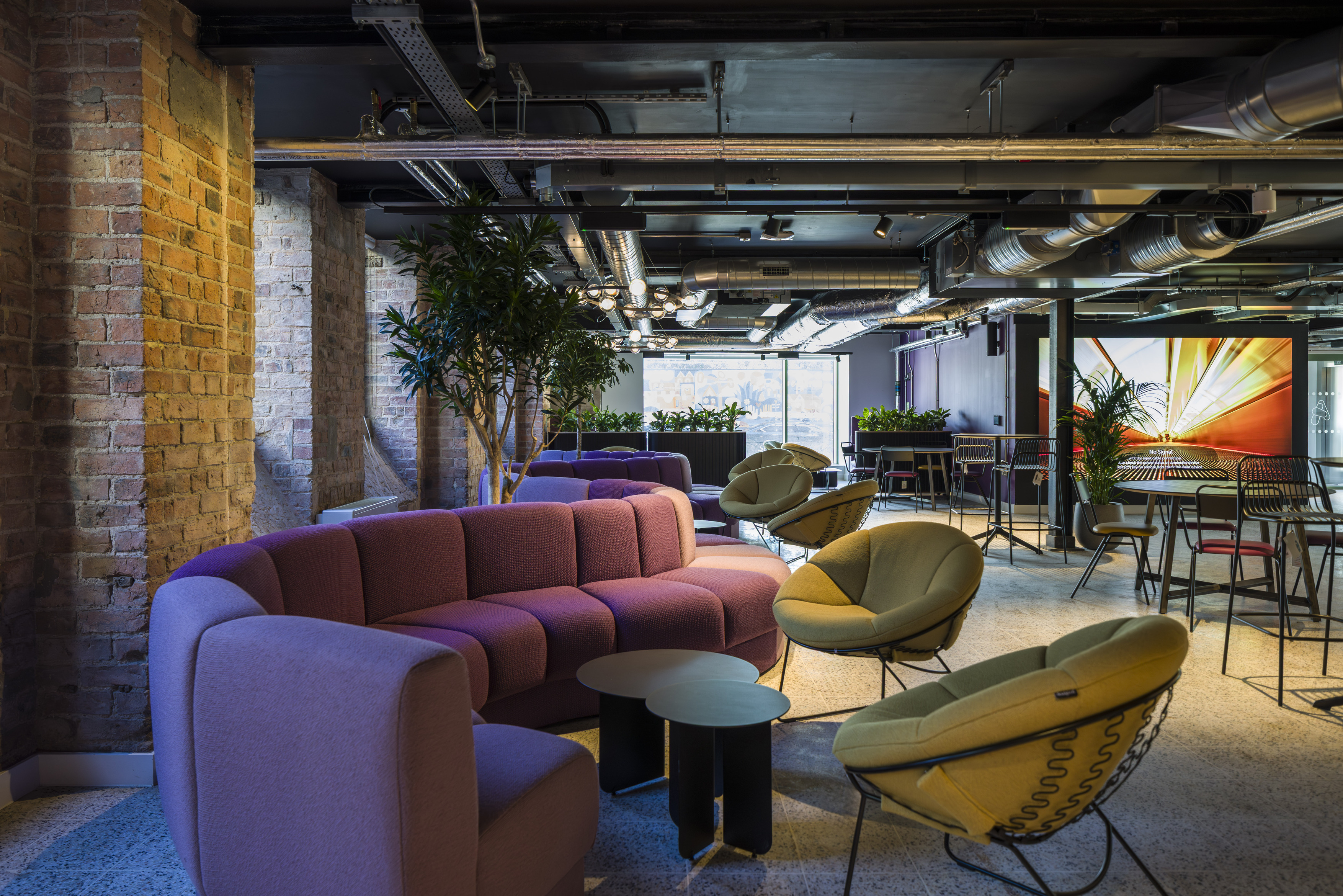 Modern office lounge with an industrial aesthetic featuring exposed brick walls and visible ductwork on a black ceiling. The space includes curved modular sofas in muted purple tones, paired with round black side tables. Two green lounge chairs with wire bases add contrast. The flooring is light terrazzo, and large potted plants provide greenery throughout. In the background, there are café-style tables and chairs, complemented by a vibrant abstract wall mural. Natural light filters through large windows, enhancing the open and collaborative atmosphere.