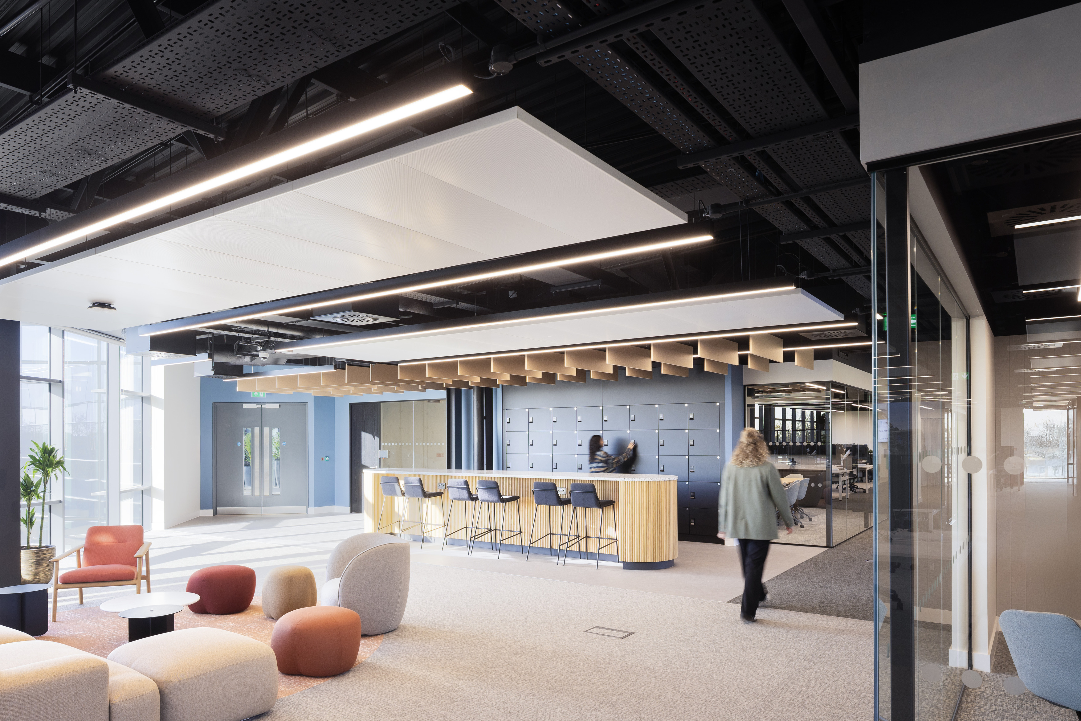 A bright open office space with soft seating in various colours arranged near full height windows. A bar height counter with stools sits in the centre of the room with a wall of lockers behind it. One person stands at the counter and another walks across the space. The ceiling features exposed black panels and linear lighting. A glass meeting room is visible on the right.