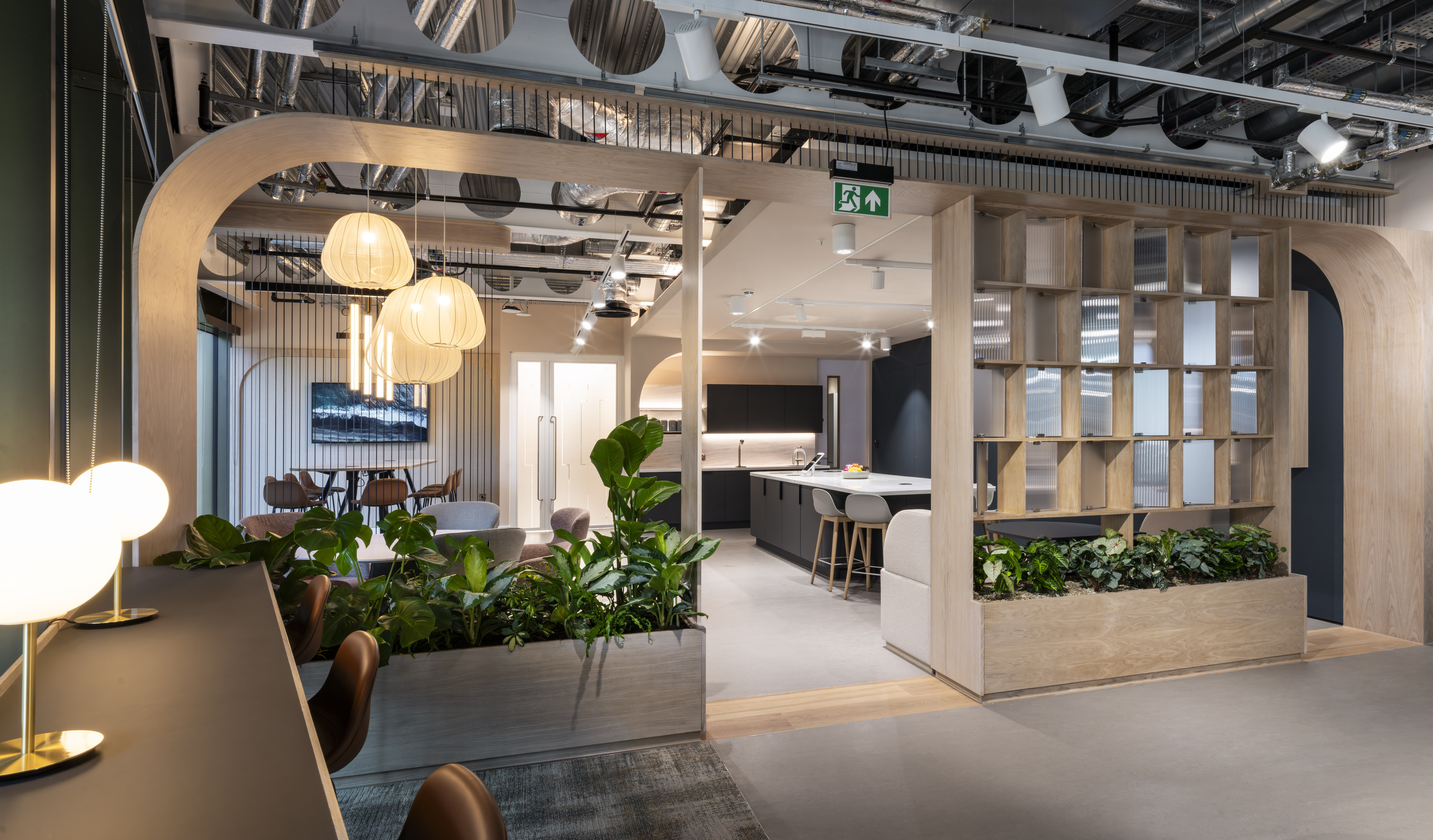 Contemporary office breakout and hospitality space featuring light oak architectural framing, integrated shelving and abundant indoor planting. The area includes lounge seating, a communal kitchen with an island and bar stools, and a separate meeting nook with a round table and pendant lighting. Exposed ceiling services, decorative acoustic panels and a mix of timber, dark cabinetry and soft furnishings create a warm, modern workplace environment with a strong biophilic design element.