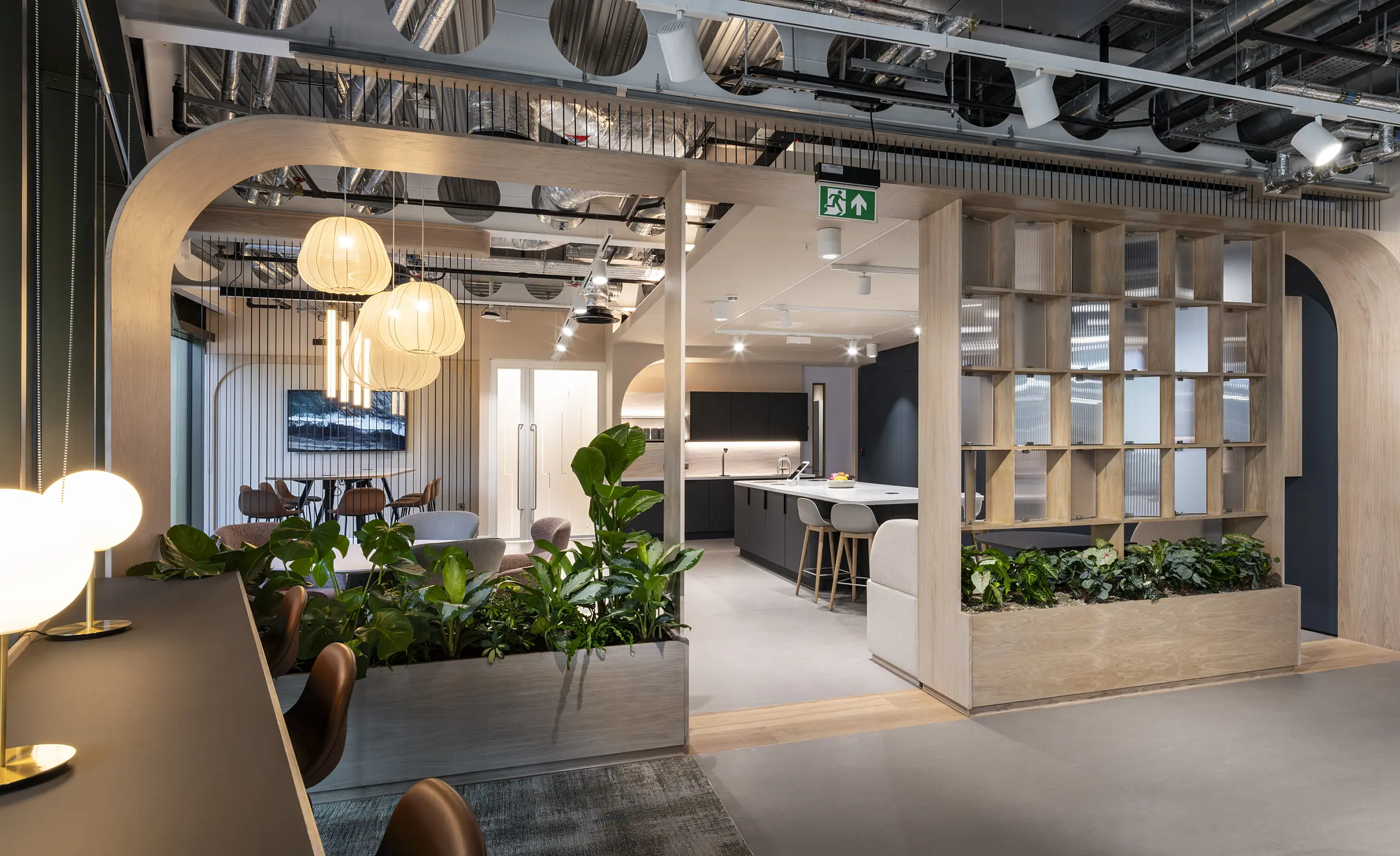 Contemporary office breakout and hospitality space featuring light oak architectural framing, integrated shelving and abundant indoor planting. The area includes lounge seating, a communal kitchen with an island and bar stools, and a separate meeting nook with a round table and pendant lighting. Exposed ceiling services, decorative acoustic panels and a mix of timber, dark cabinetry and soft furnishings create a warm, modern workplace environment with a strong biophilic design element.