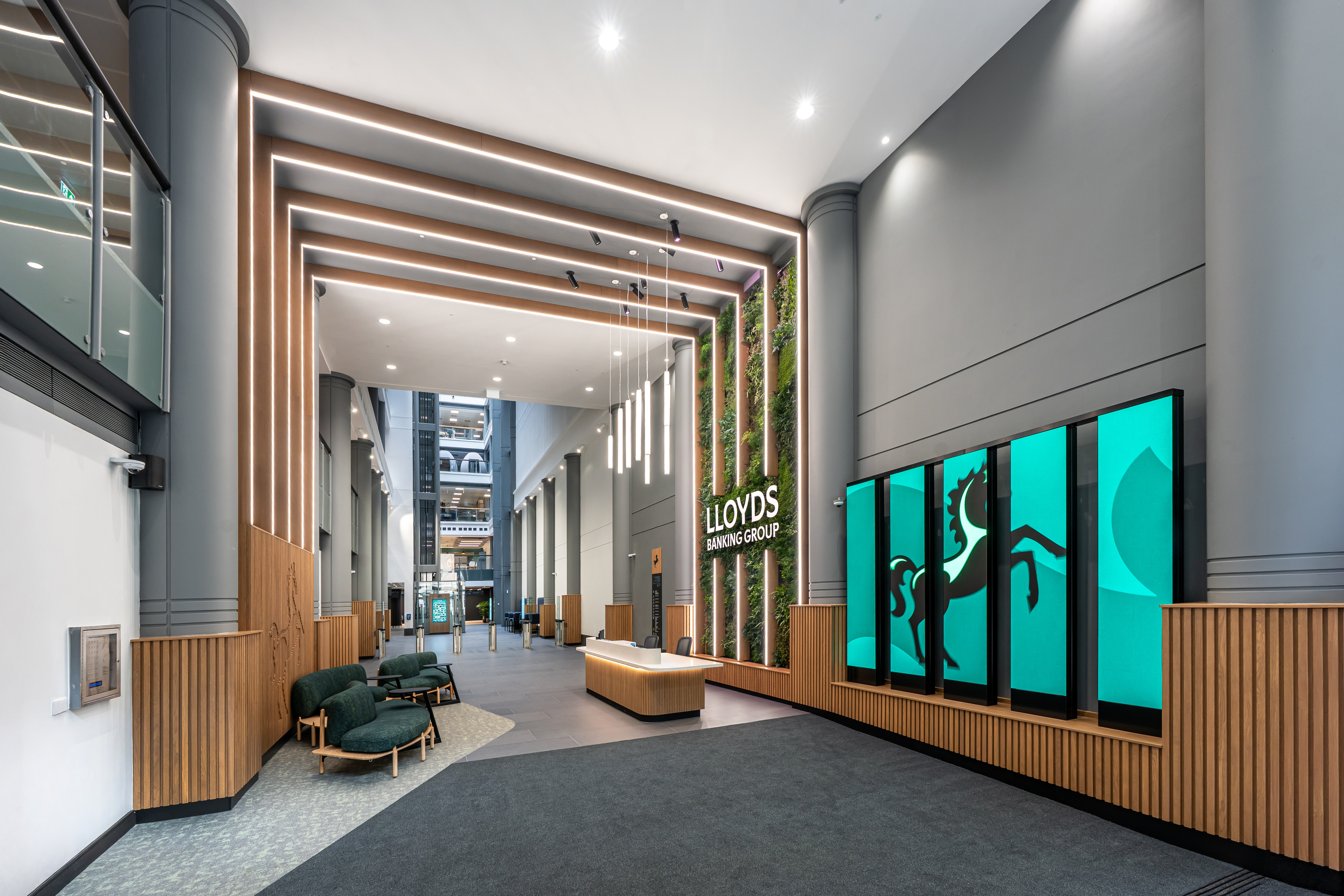 A spacious modern lobby with high ceilings and tall grey walls featuring vertical wooden slats. A series of suspended linear lights hangs above a central reception desk made of light wood. To the right, a green living wall surrounds the Lloyds Banking Group logo. Next to it, large digital screens display the company’s black horse symbol on a teal background. Soft seating with dark green upholstery sits to the left, and the far end of the space opens into a bright atrium with additional architectural details.