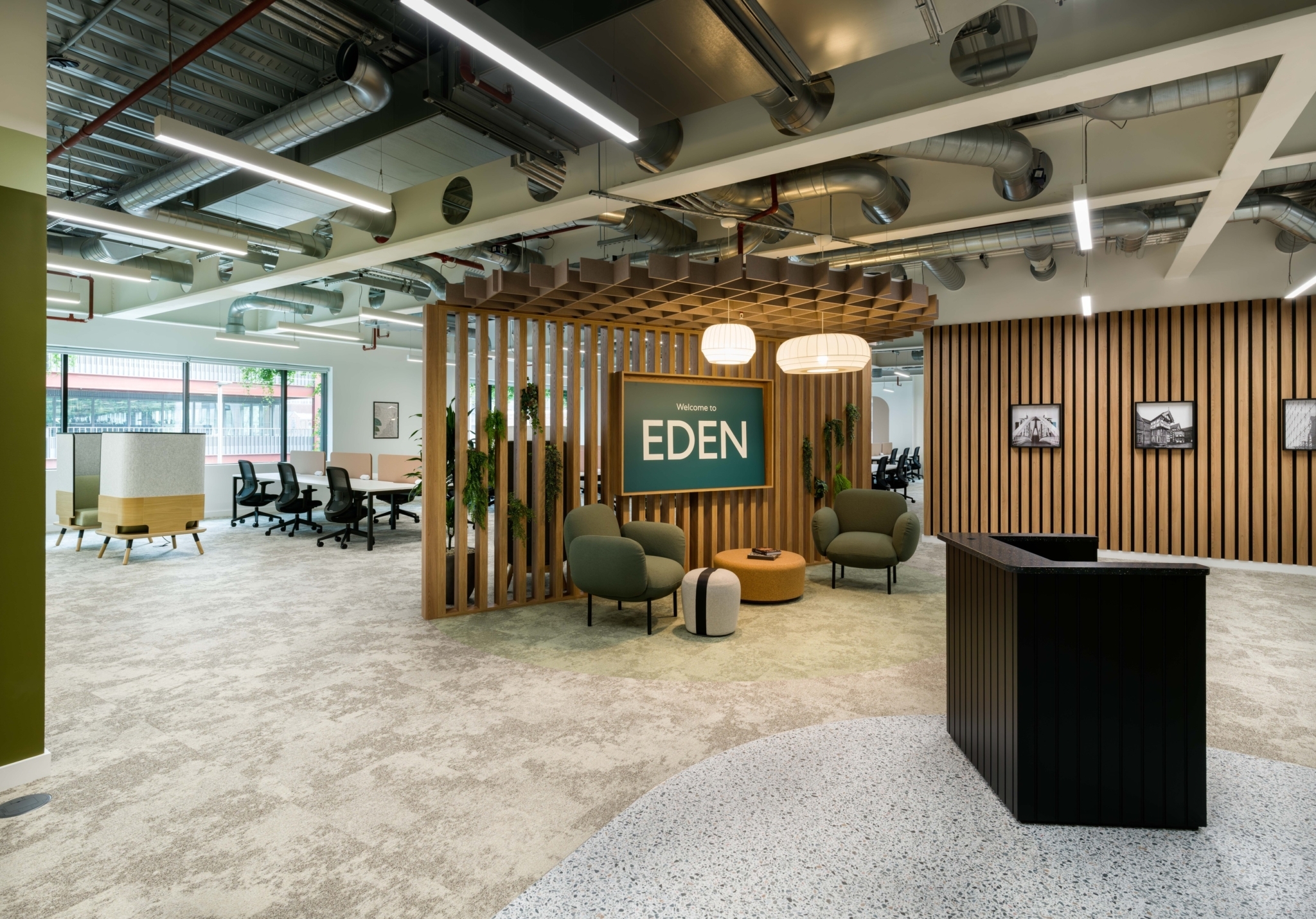 Bright, modern office reception area with an open-plan layout. In the centre, a wooden slatted feature structure frames a green sign reading “Welcome to EDEN”, with soft seating including armchairs and a round ottoman arranged beneath warm pendant lights. To the right, a dark reception desk sits against a wall of vertical wooden slats displaying framed black-and-white photos. To the left, a workspace with desks and office chairs is visible through large windows. The ceiling features exposed ductwork, pipes and linear lighting, while the floor combines carpet and polished concrete finishes, giving the space a contemporary, industrial feel.
