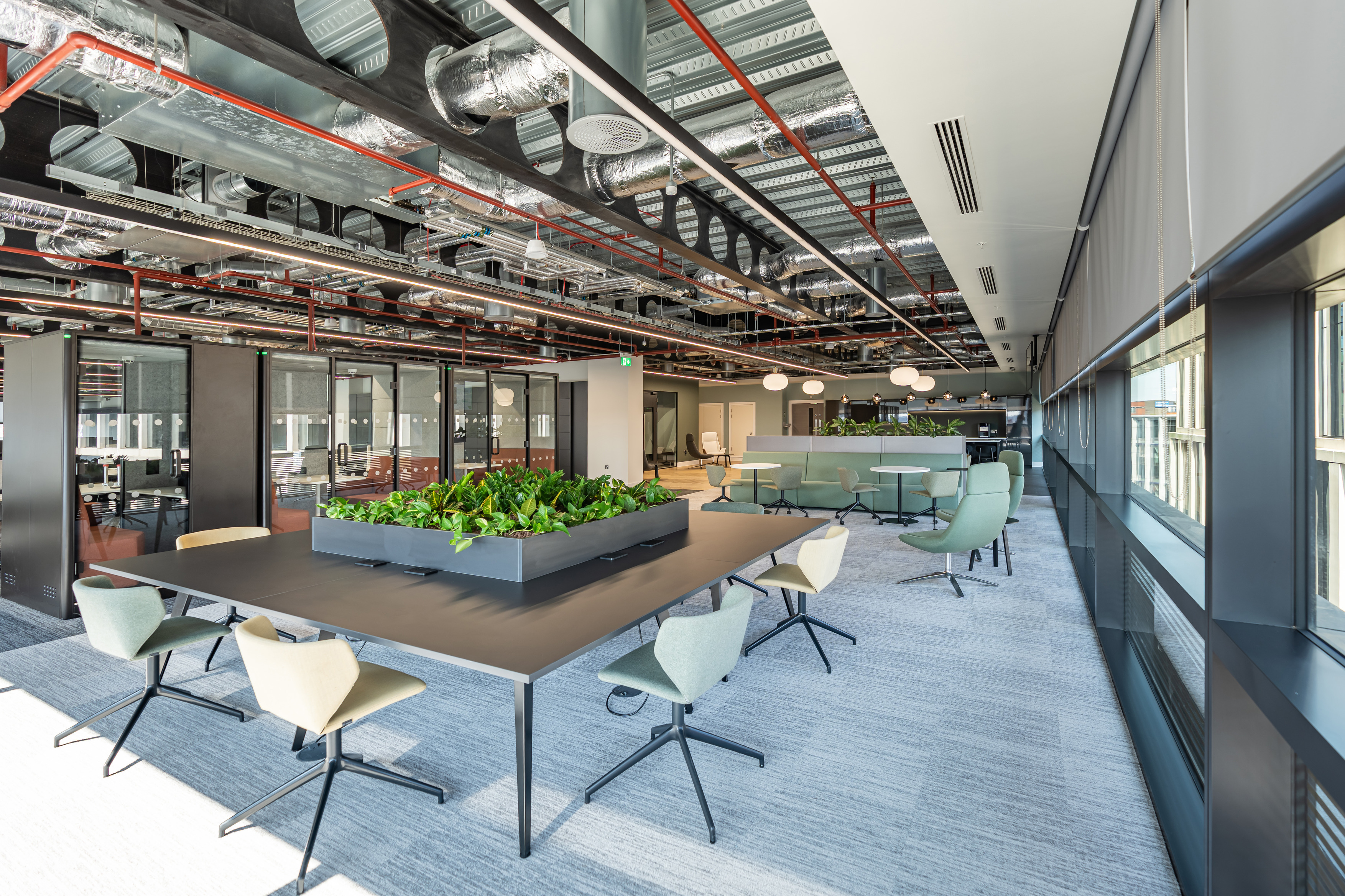 A modern open plan office with an industrial aesthetic featuring exposed ceilings with visible ductwork and piping. Large windows on the right side let in natural light. A long rectangular table with a central planter box filled with green plants sits in the middle, surrounded by swivel chairs in muted green and beige. Toward the back, casual meeting areas include round tables and comfortable chairs. The floor is covered in light grey carpet tiles, contributing to the contemporary look.